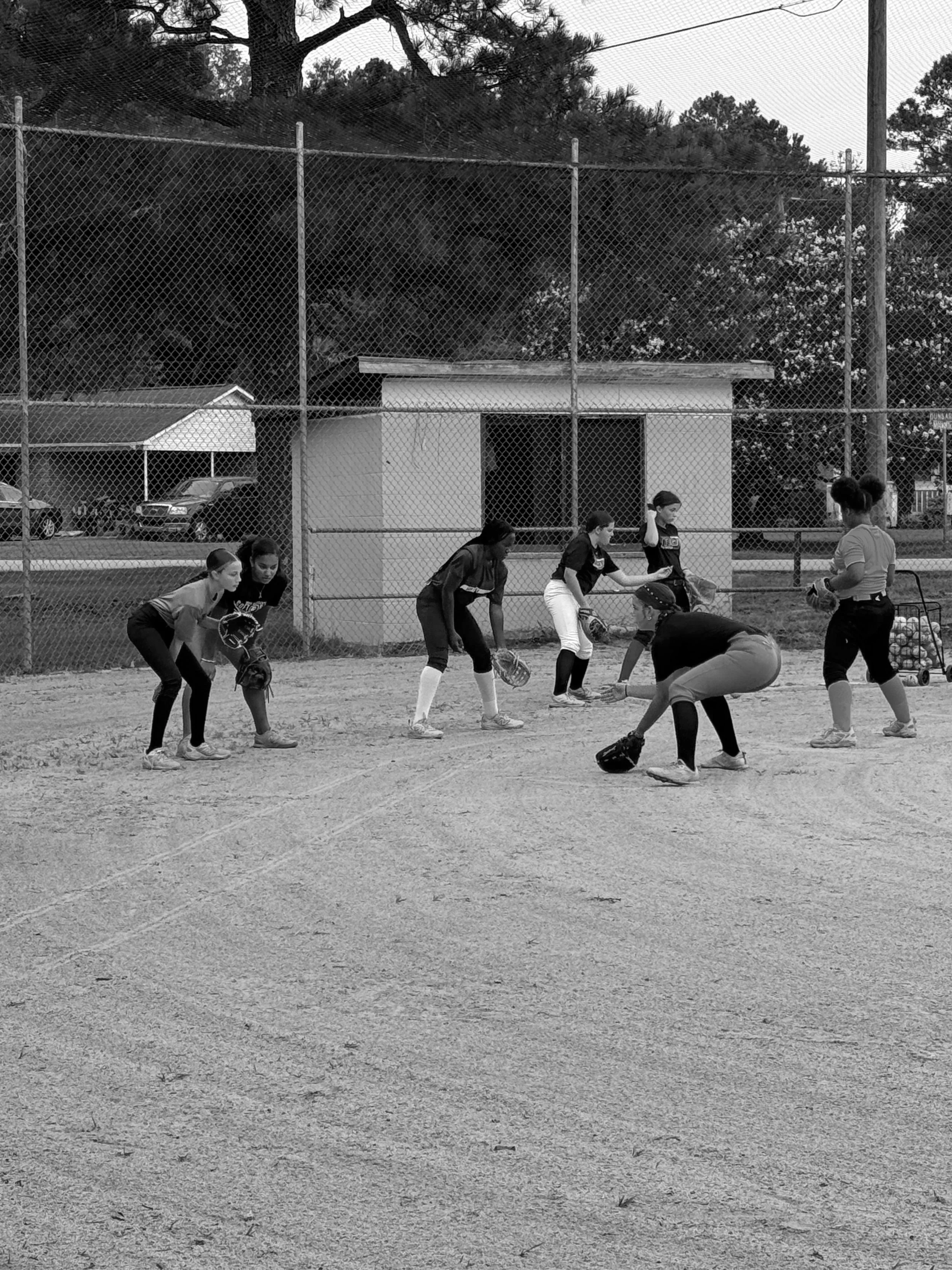 A group of young female softball players in uniforms practicing in a dirt field, with a chain-link fence, trees, and parked cars in the background.