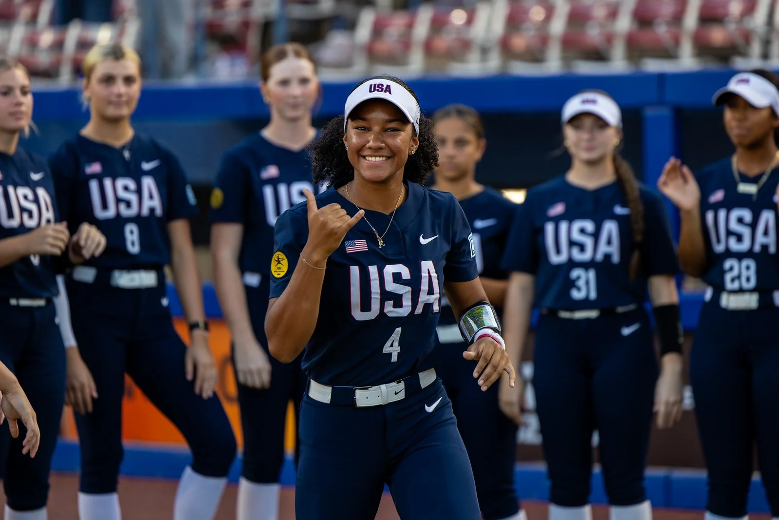 A group of female athletes in USA uniforms, with one in the center making a shaka sign with her hand, smiling, on a sports field.