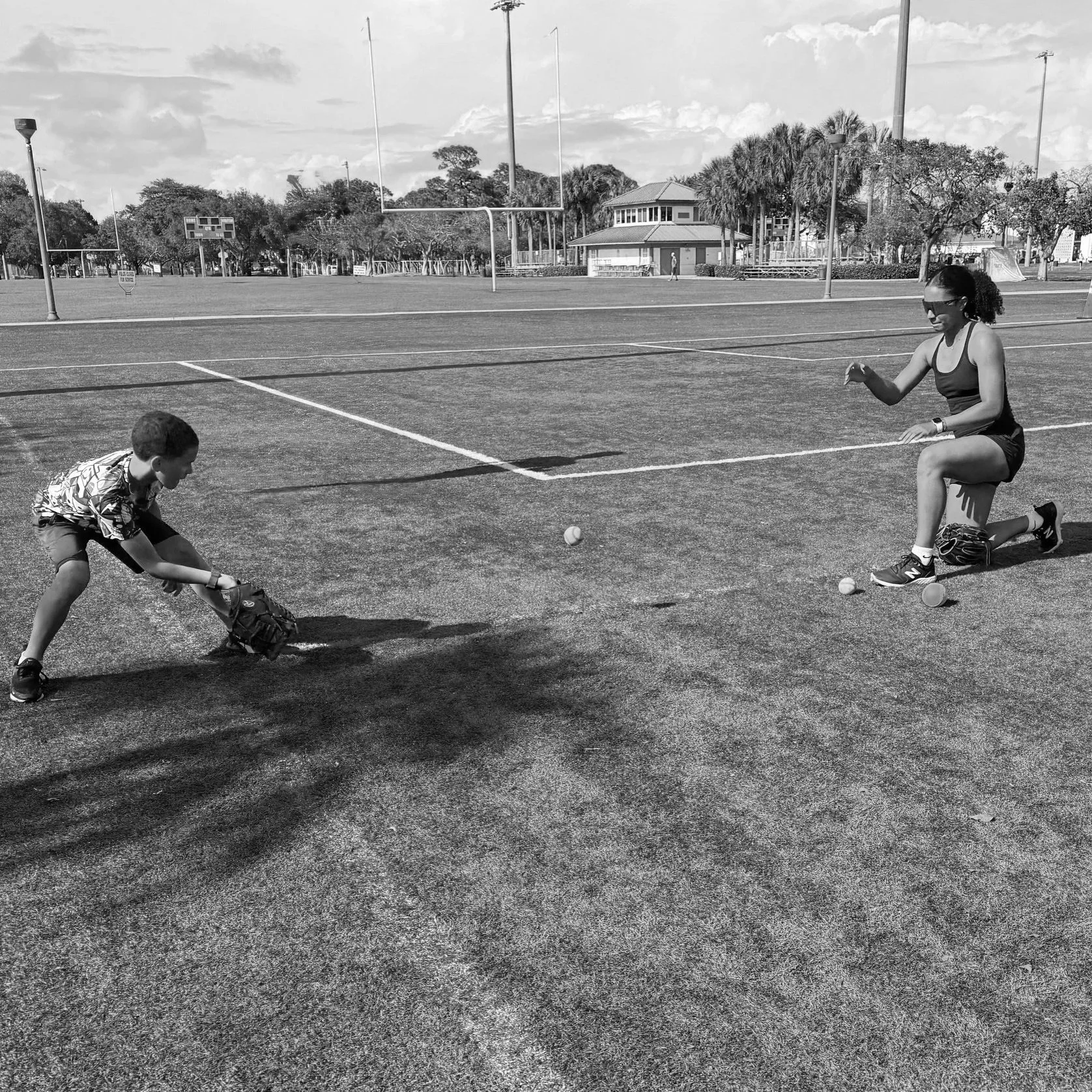 A woman and a boy playing catch with baseballs on a grassy field with bleachers and trees in the background.