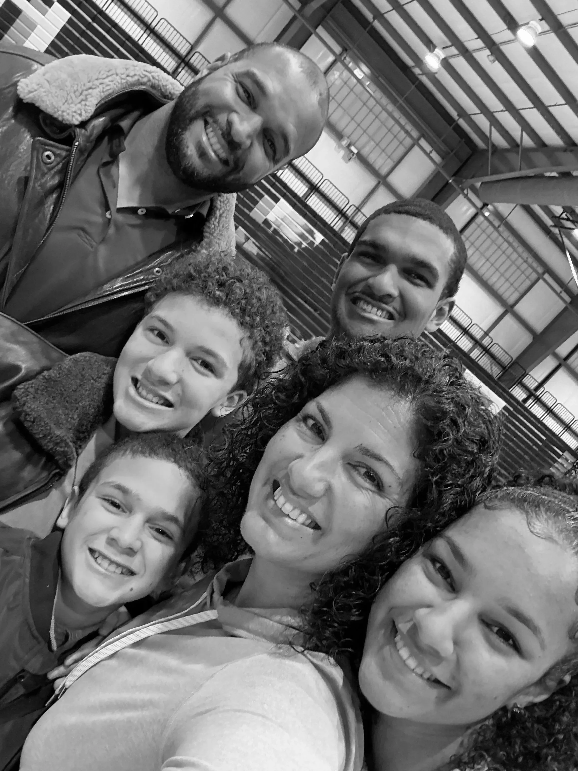 Group of smiling people taking a selfie in an indoor setting with scaffolding or metal structures in the background. The image is in black and white.