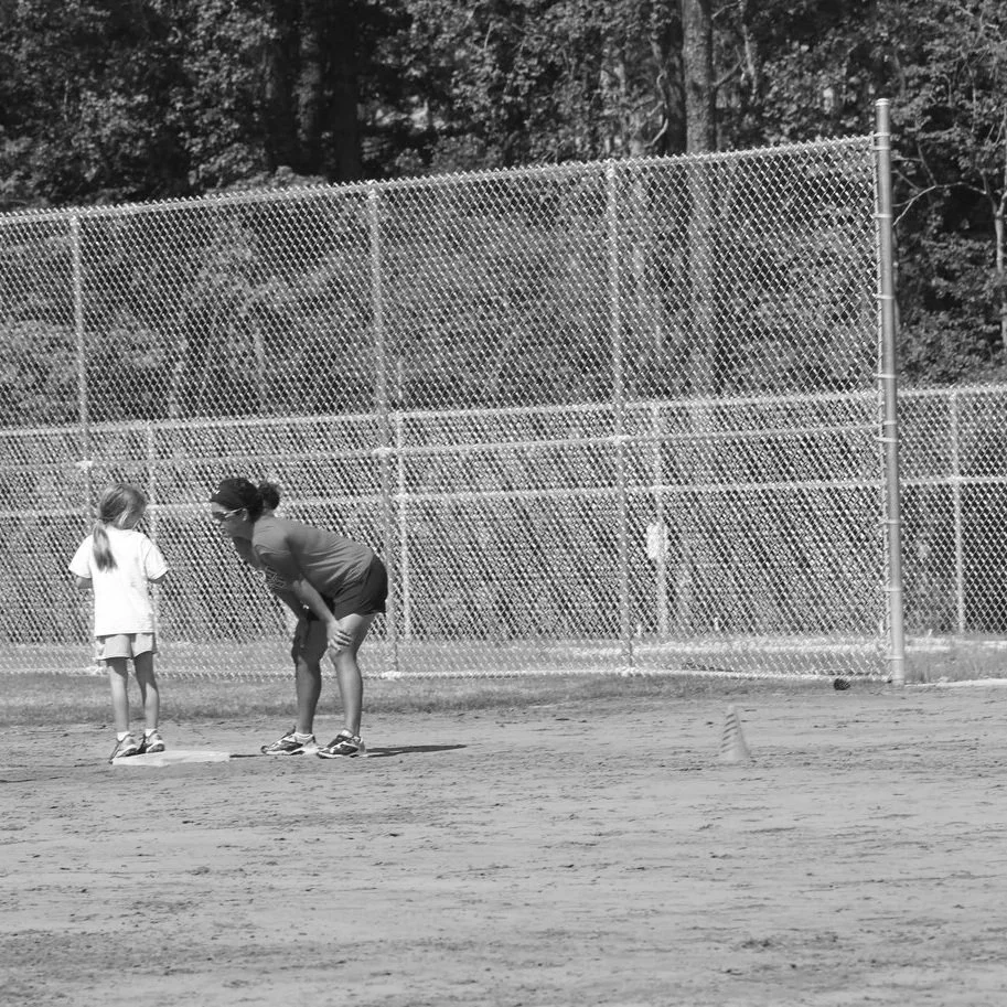 A woman and a young girl are on a baseball field, suggesting they are preparing for or playing a game, with a tall chain-link fence in the background.