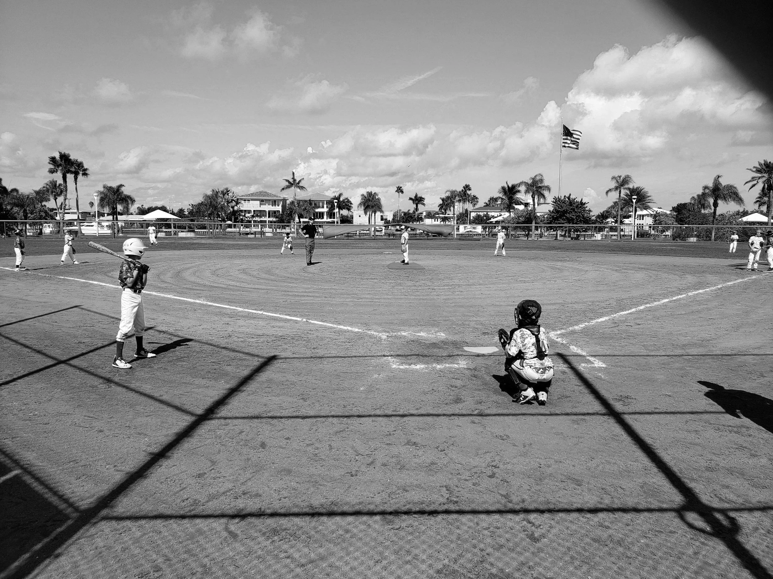 Black and white photo of a youth baseball game at a park, with players on the field, a coach, and a bat boy holding a bat. There is a chain-link fence in the foreground and houses and palm trees in the background under a partly cloudy sky.