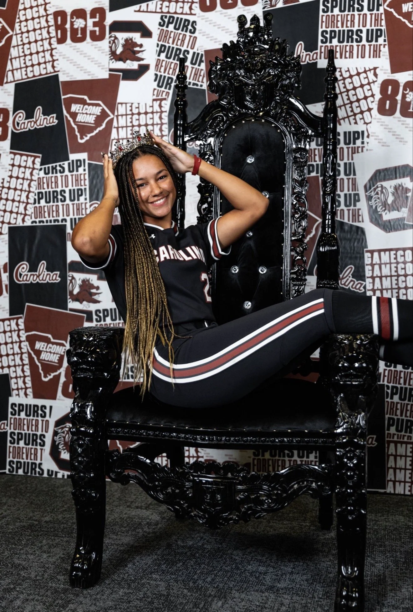 Young girl sitting on a black, ornate throne-like chair, wearing a jersey and a crown, smiling, with sports and team logos and slogans in the background.