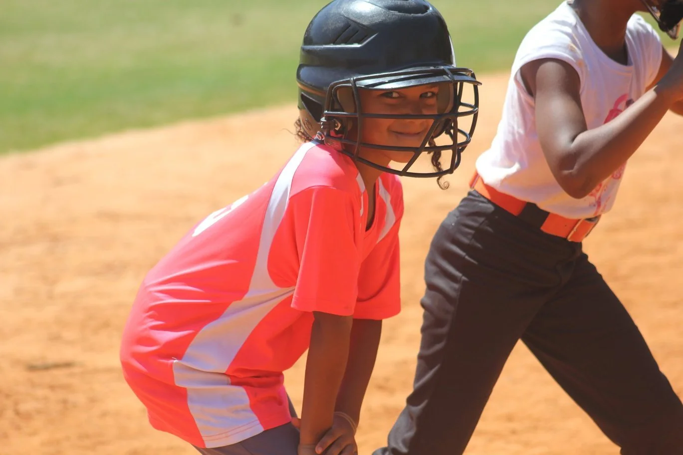 Young girl wearing a black baseball helmet, pink and white sports uniform, smiling, on a baseball field.