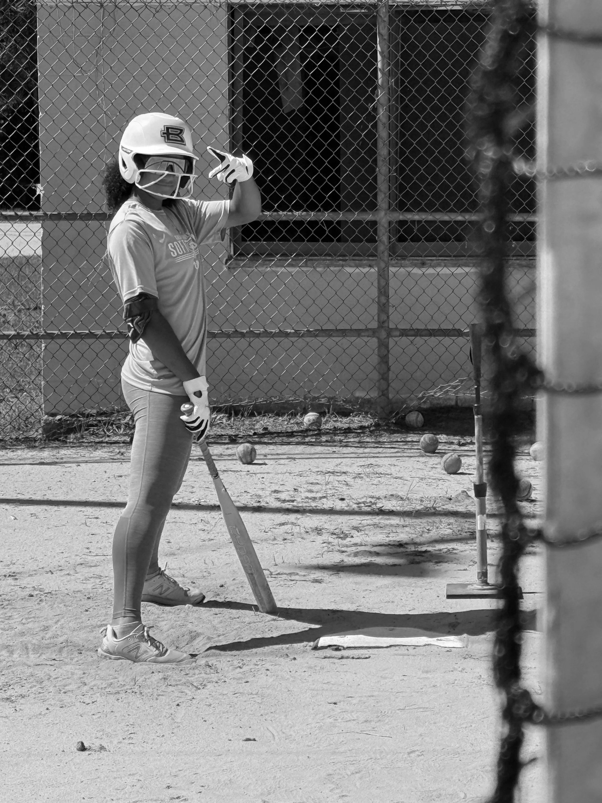 A person wearing a baseball helmet and gloves standing on a baseball field, preparing to catch or hit a ball. There are balls scattered on the ground nearby, and a baseball net or fence is in the foreground.