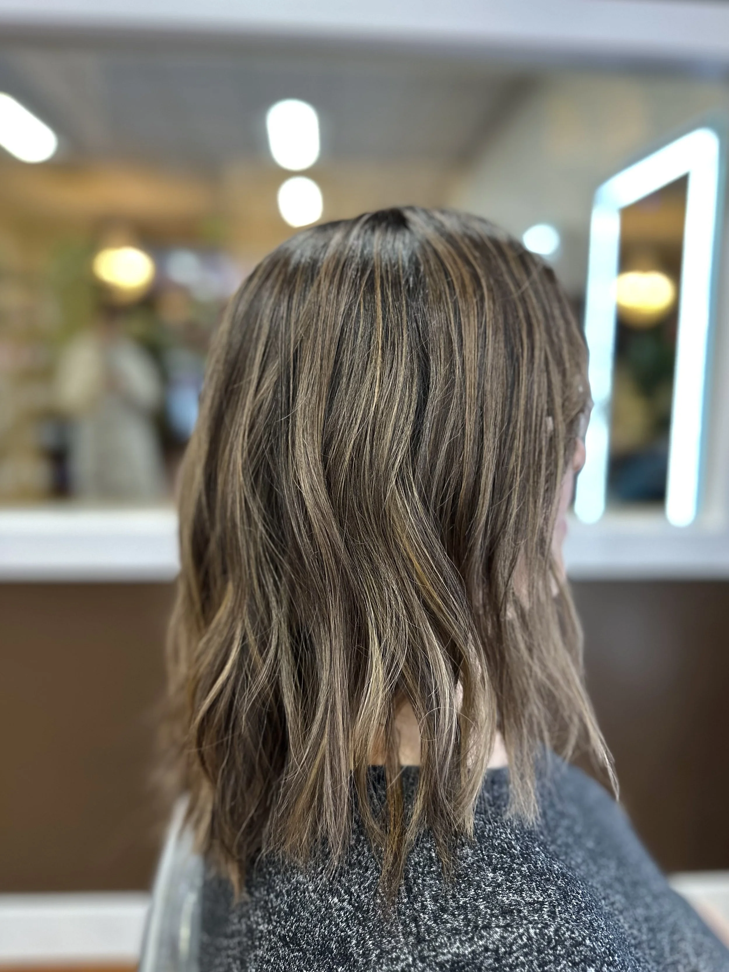 A person with long, wavy, brown hair with blonde highlights, wearing a black and grey spotted top, is looking at a mirror in a salon or hair studio.