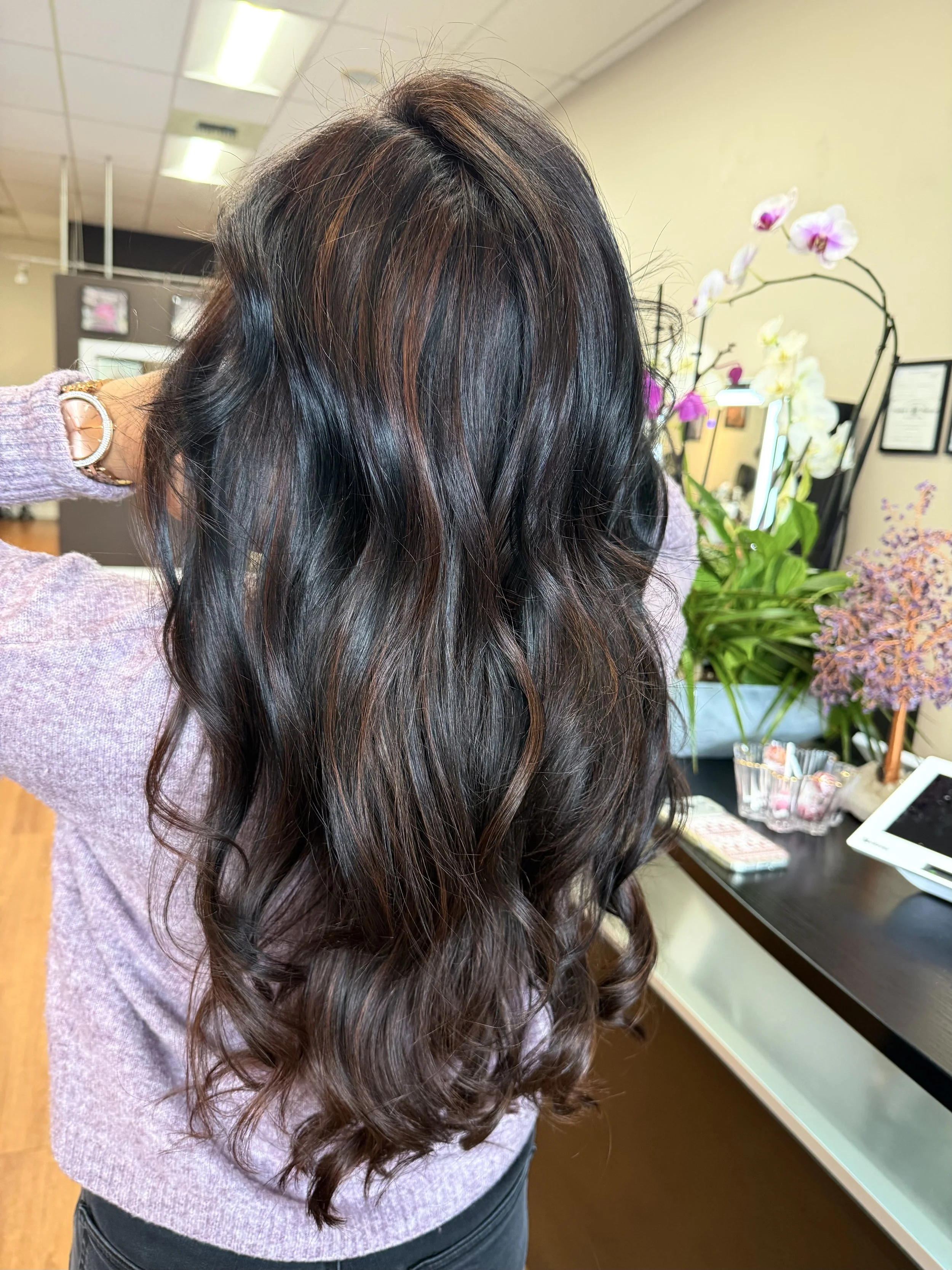 A woman with long, dark, wavy hair with brown highlights standing in a room with flower arrangements on a table and framed pictures on the wall.