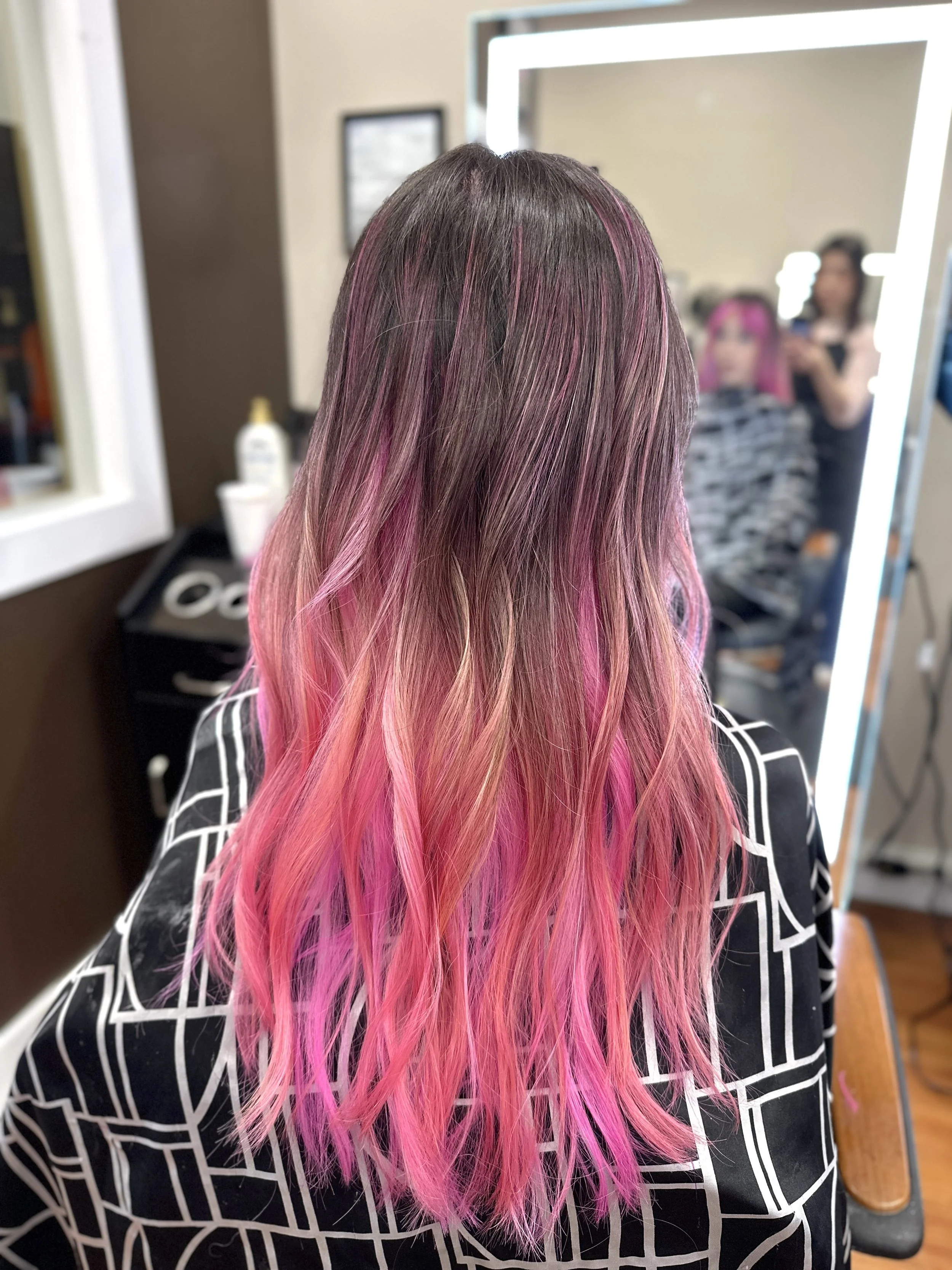 Back view of a woman with long hair, dark roots, and pink highlights, sitting in a salon chair in front of a mirror, with a stylist and a client visible in the reflection.