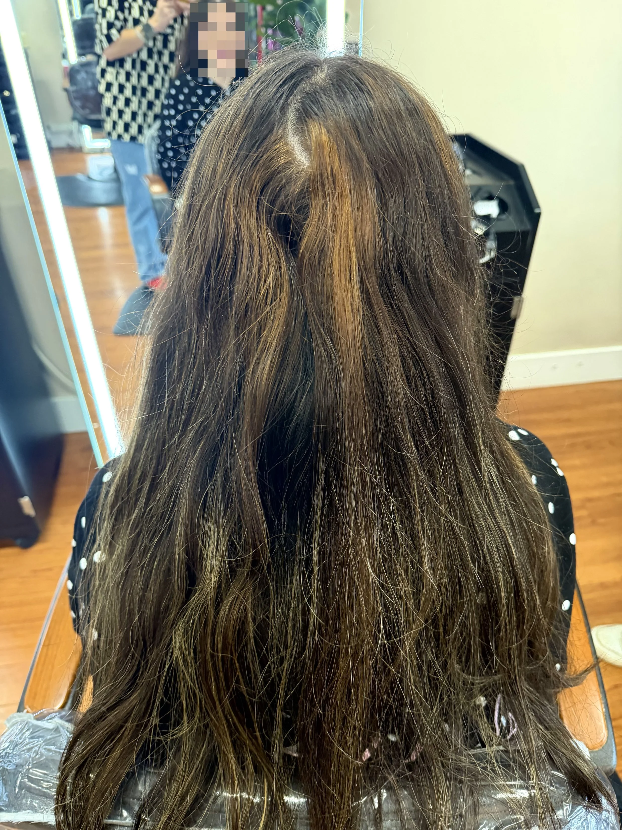 A person with long brown hair sitting in a salon chair, facing away from the mirror, with hair sectioned and prepared for a hair treatment or styling. Hairdresser and customer are visible in the background.