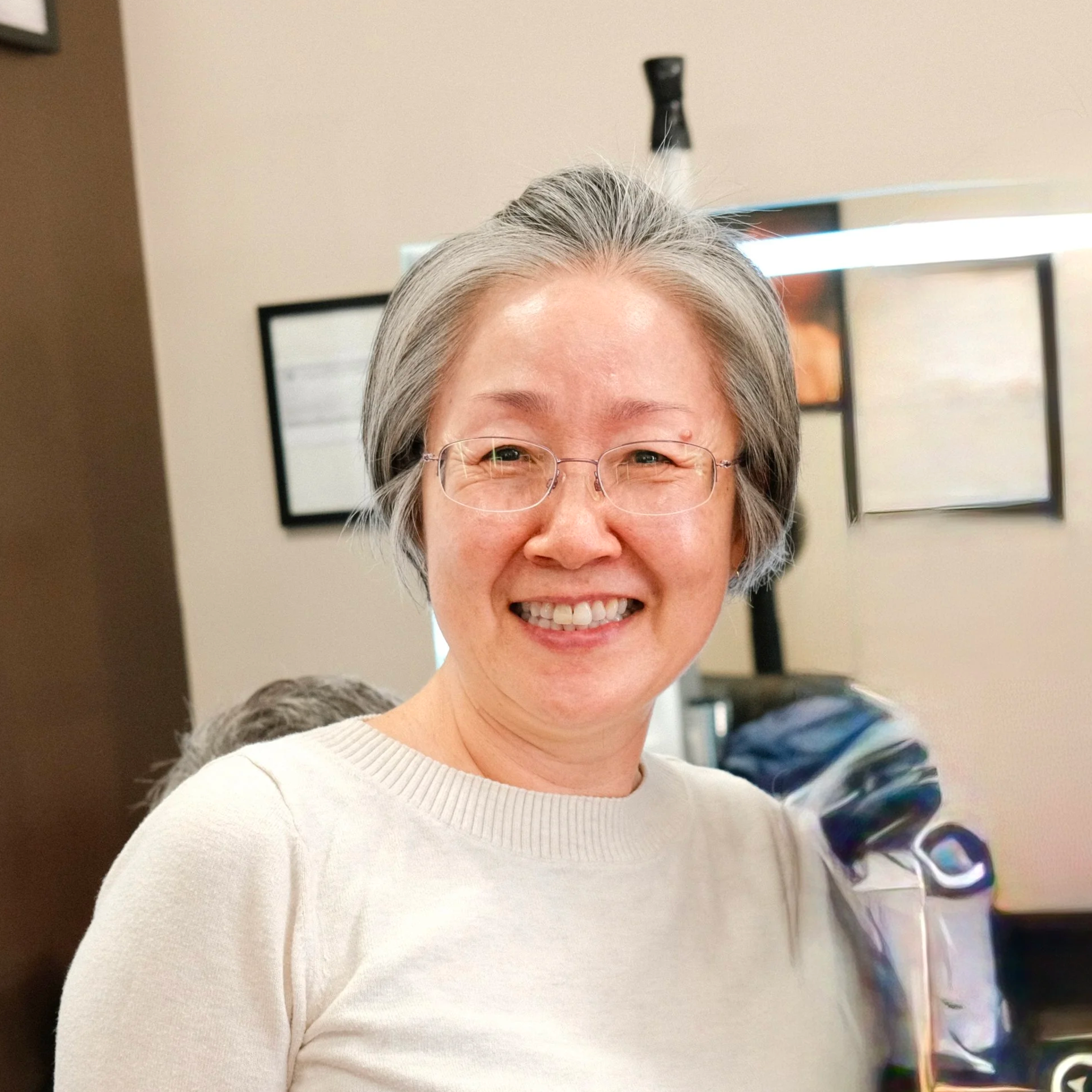 Close-up of an elderly woman with gray hair wearing glasses, smiling, indoors with a beige wall and a computer monitor in the background.