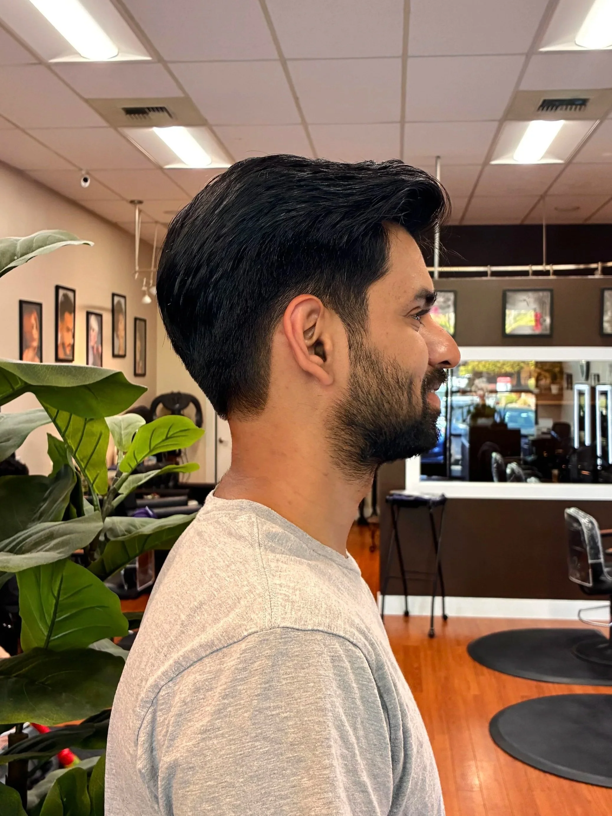Side profile of a man with dark, well-groomed hair and a beard inside a hair salon or barbershop.