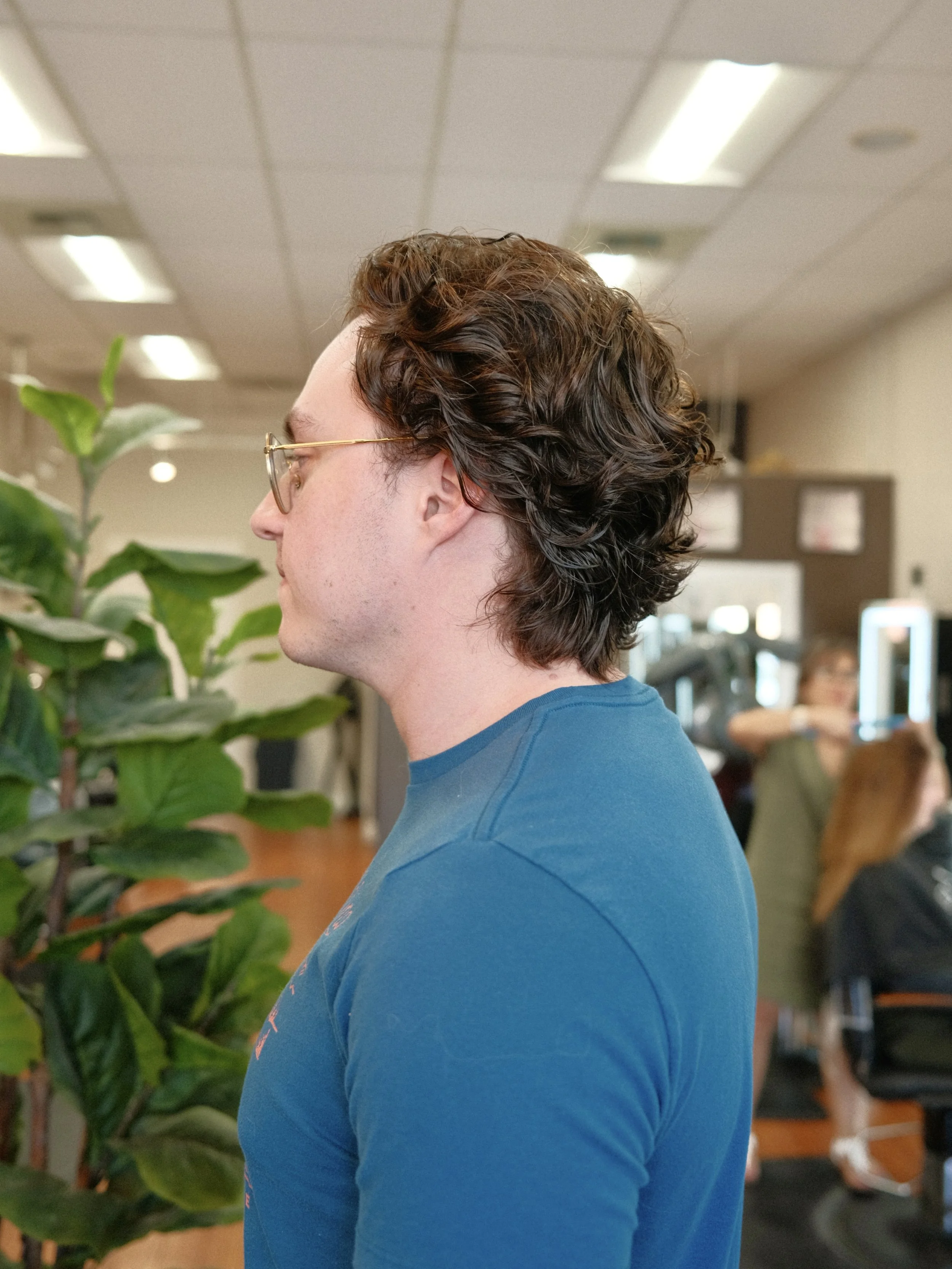 Profile of a young man with curly brown hair, wearing glasses and a blue T-shirt, standing indoors near a green plant, with a blurred background showing people and an office environment.