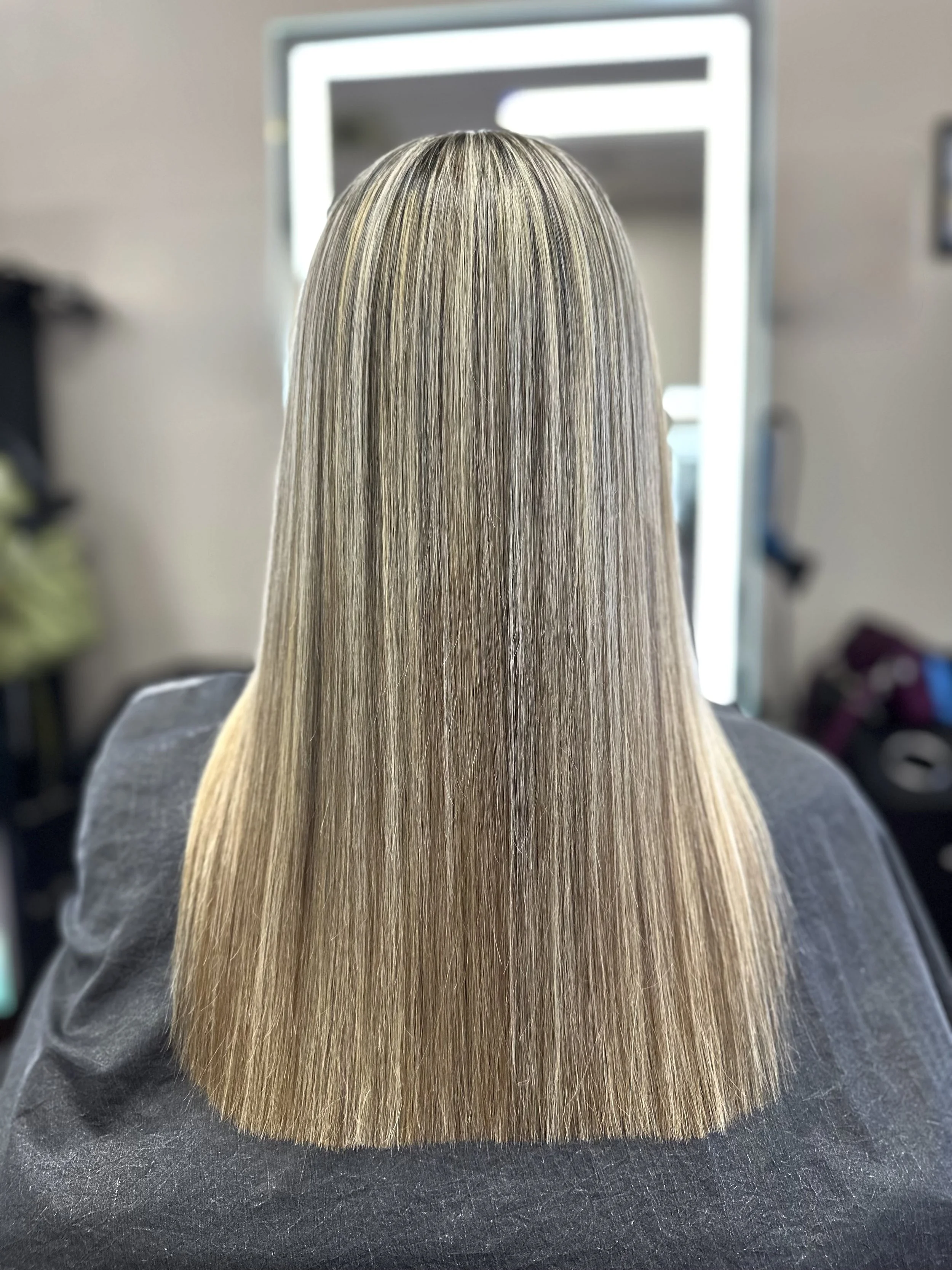 A person with straight, long blonde hair sitting in front of a mirror in a salon.