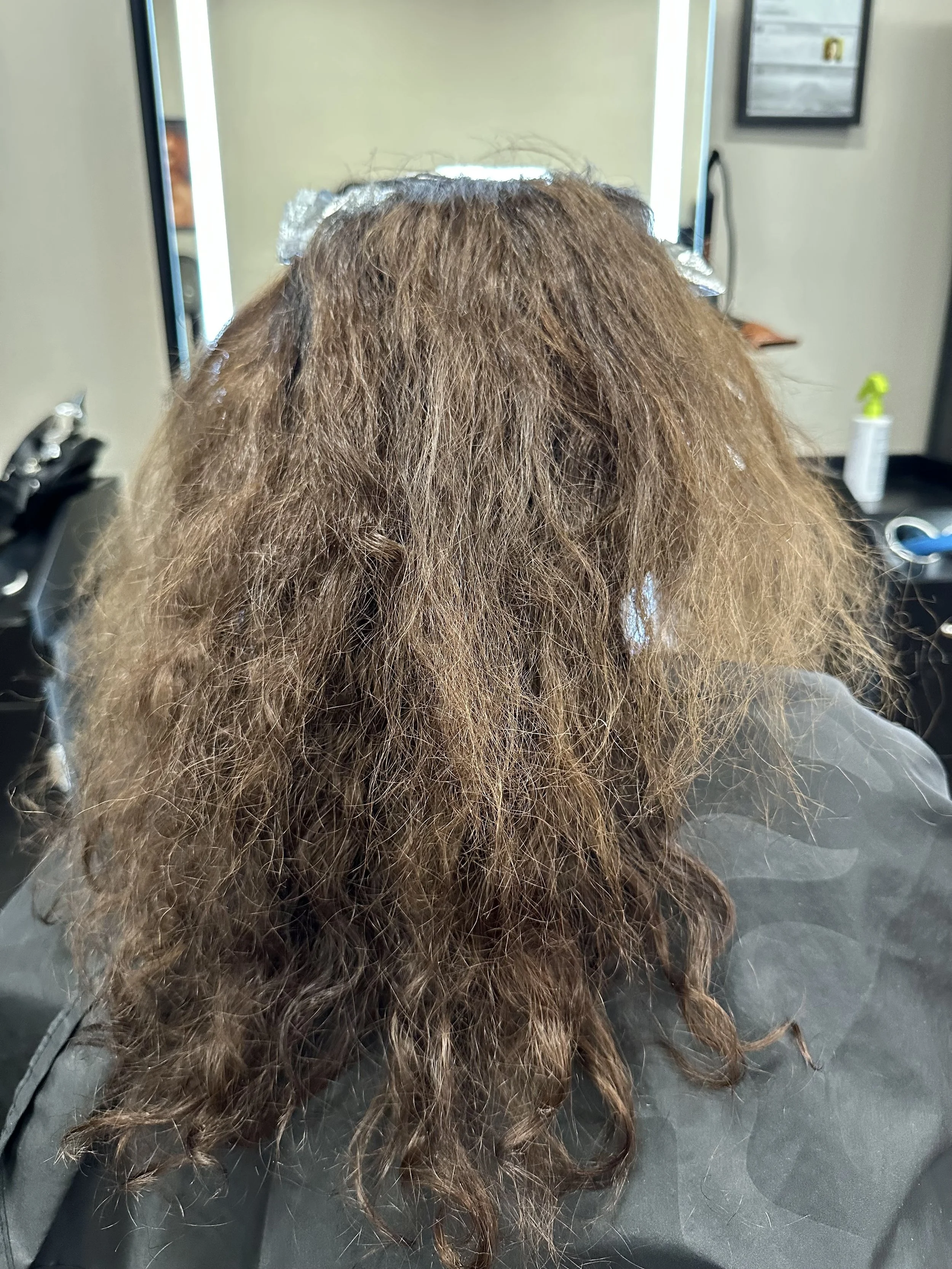 A person with light brown, curly hair sits at a salon chair with a foil hair treatment on top of their head, in front of a mirror.