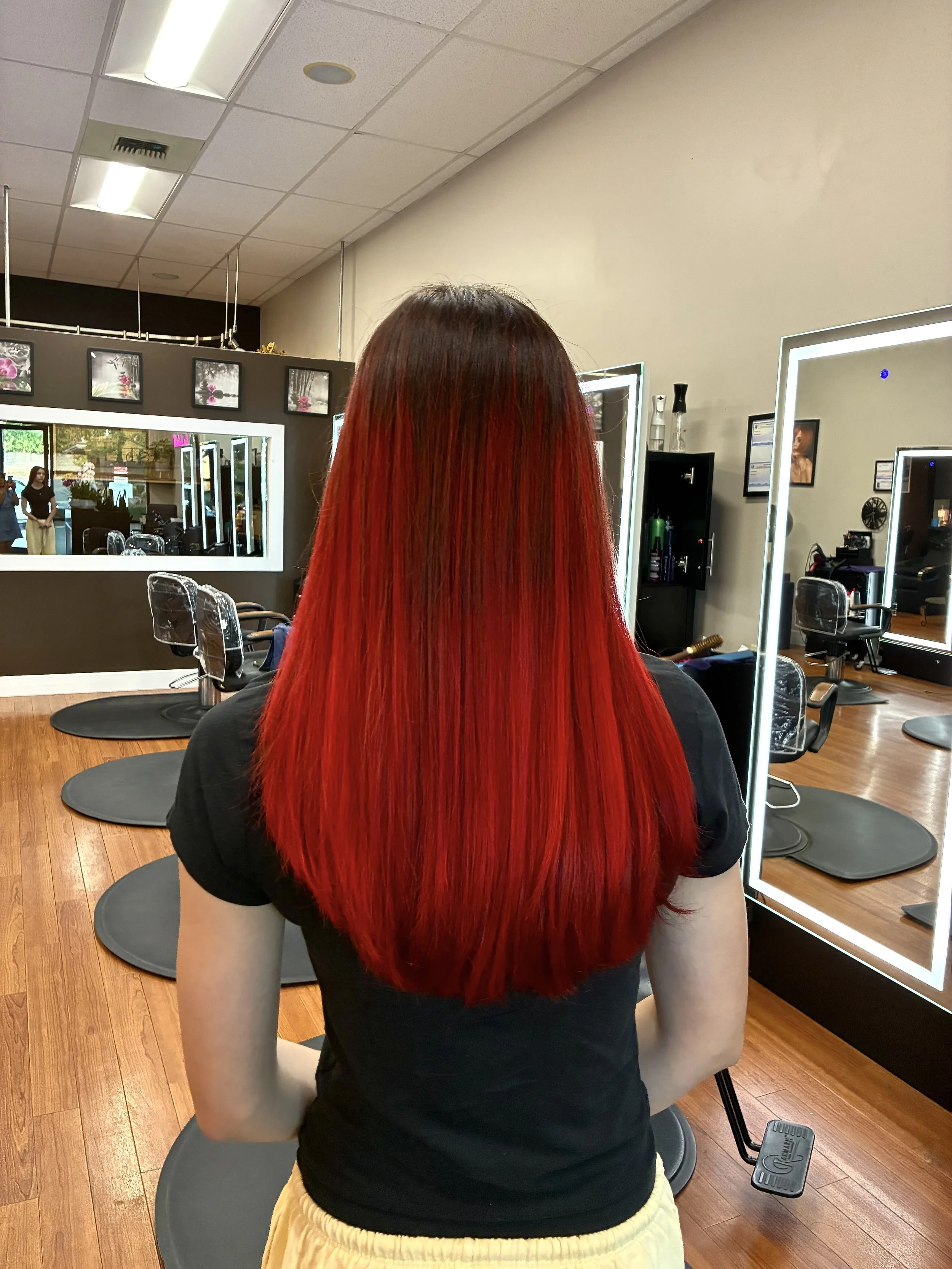 Person with long red hair sitting in a salon chair, facing away from the camera, with mirrors and salon equipment visible in the background.