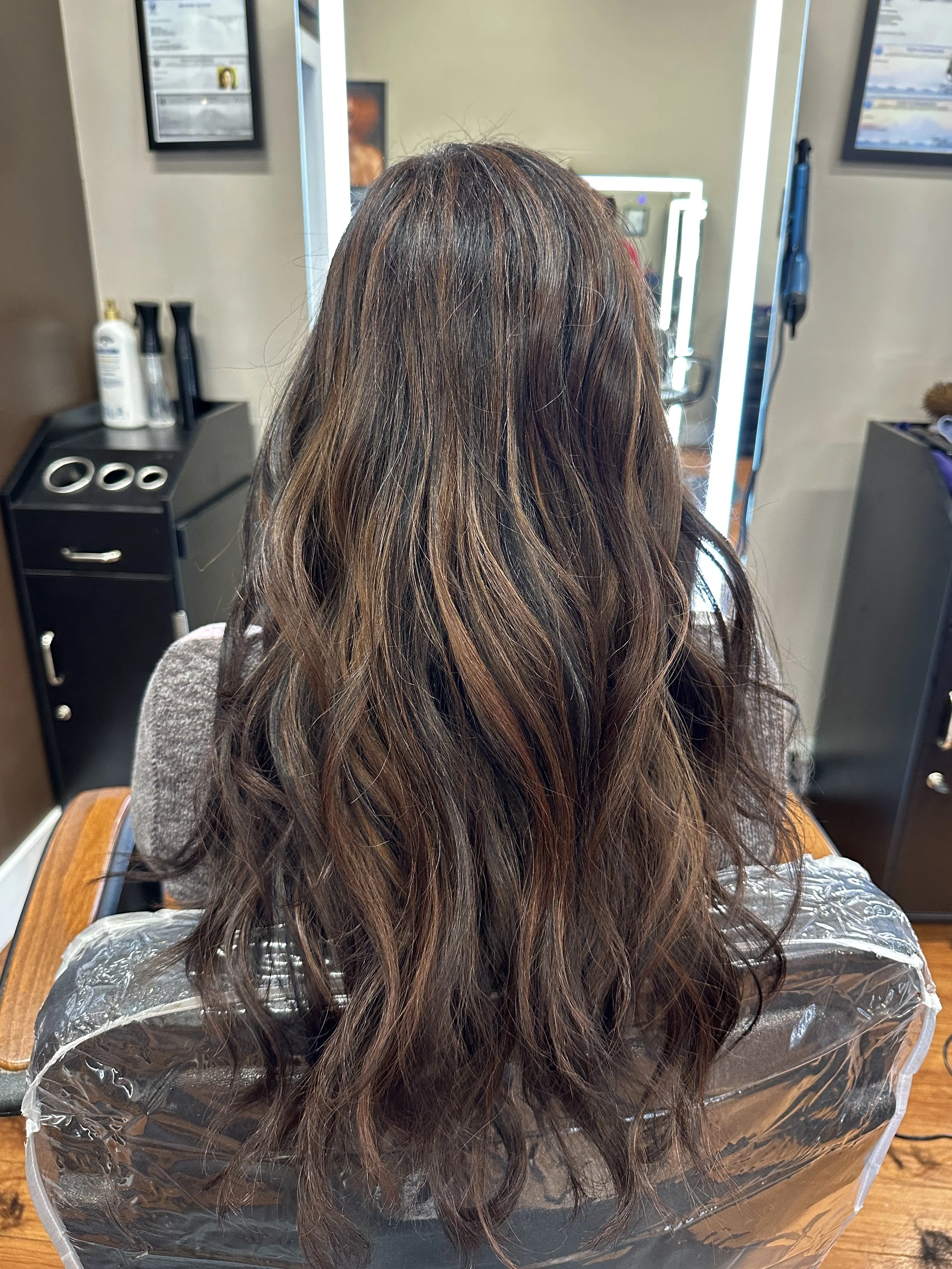 The image shows the back of a woman with long, wavy, brown hair, sitting in a salon chair covered with a plastic cape. The salon interior with a mirror and hair products is visible in the background.