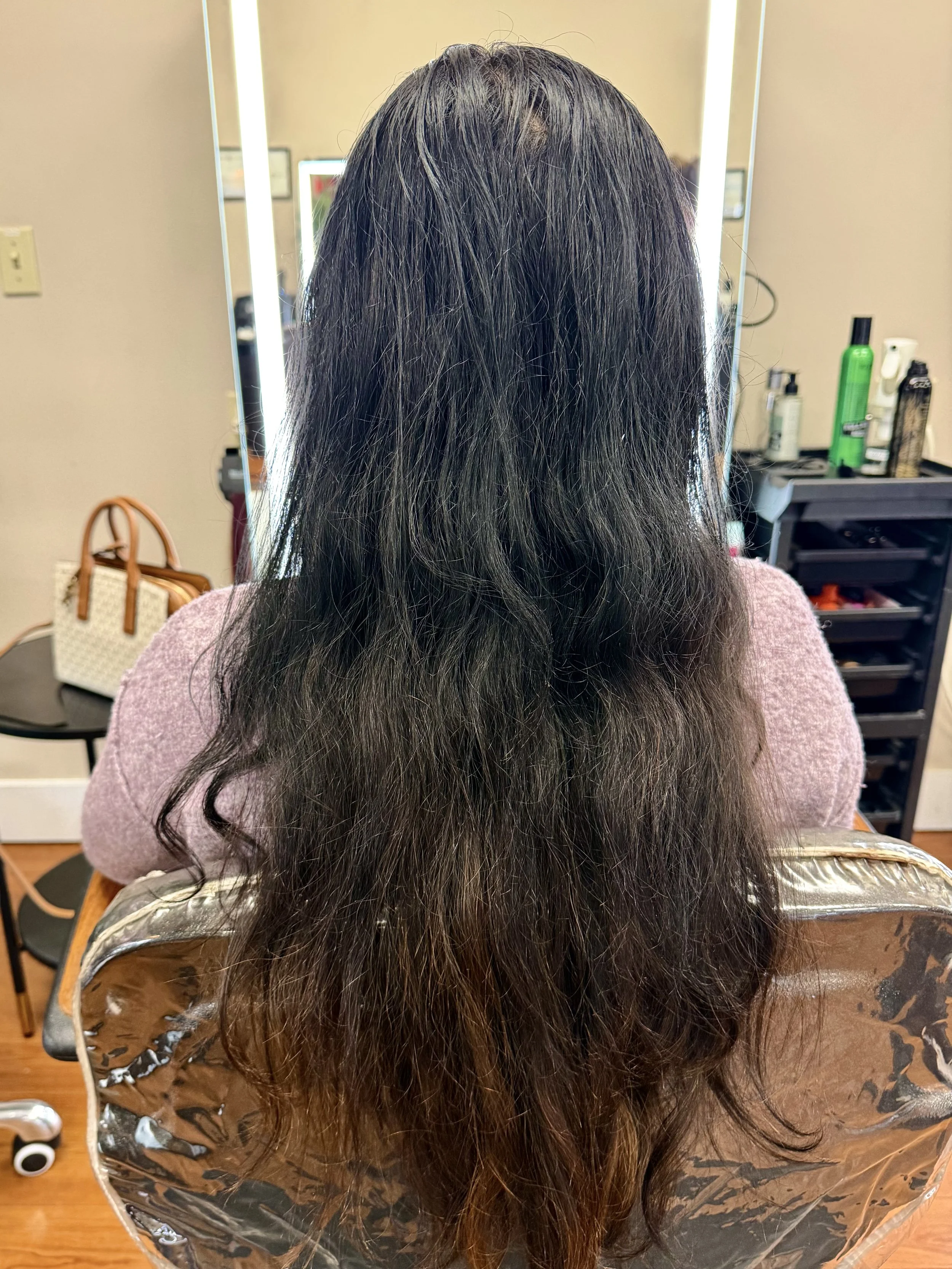 The image shows dark brown and black hair falling over a person's back, sitting in a salon seat with a reflective mirror in front. The background includes salon products on a shelf and a black rolling cart.