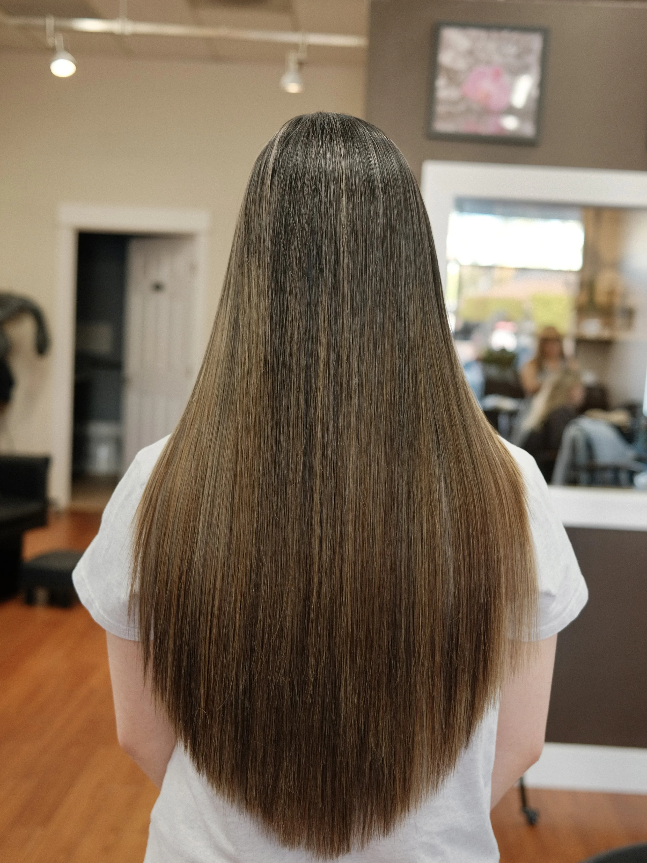Back of a woman's head with long, straight, brown hair, in a hair salon with a mirror and other salon chairs in the background.