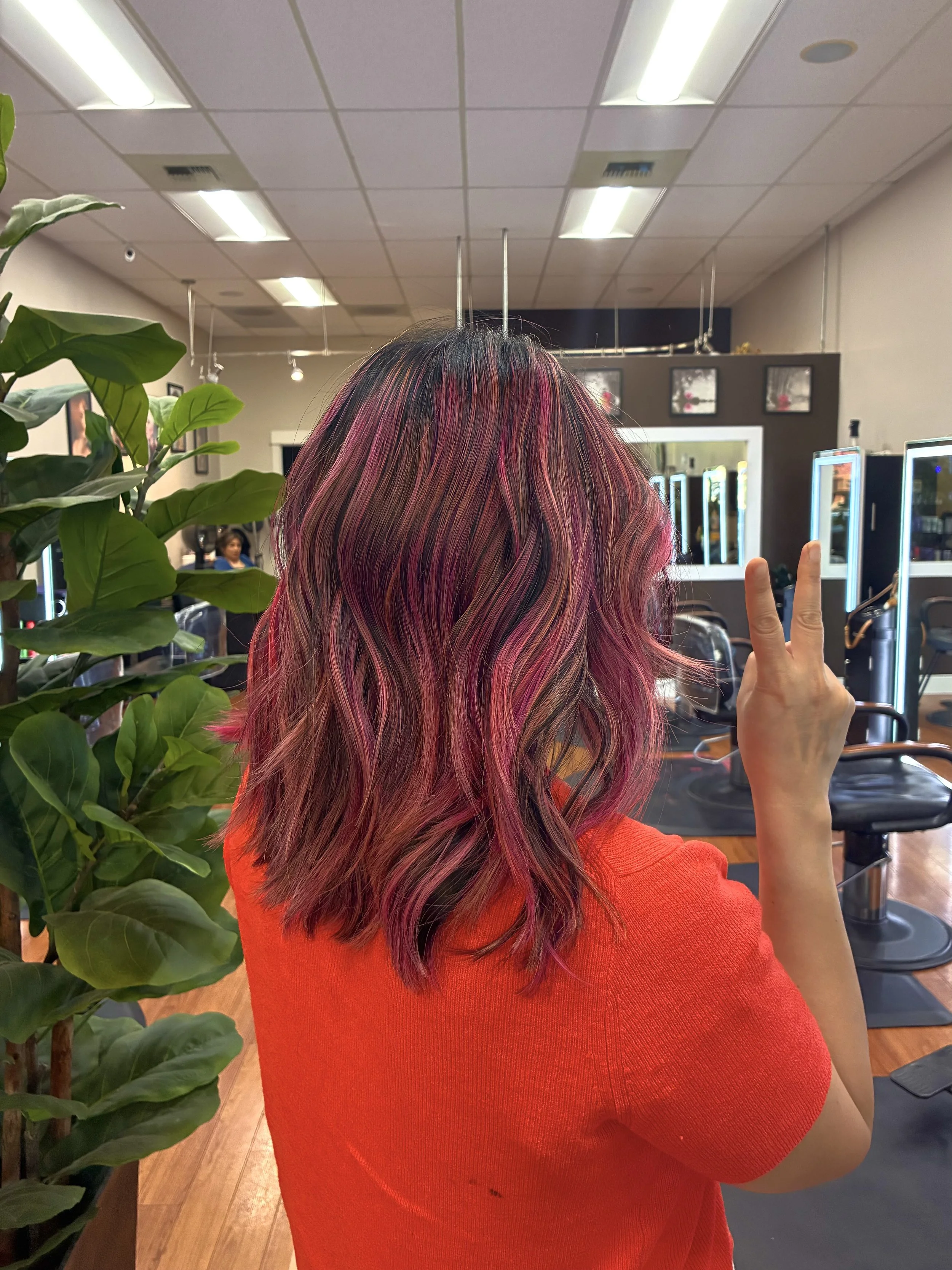 A person with long hair dyed in shades of pink, red, and black, showing a peace sign with their right hand in a hair salon.