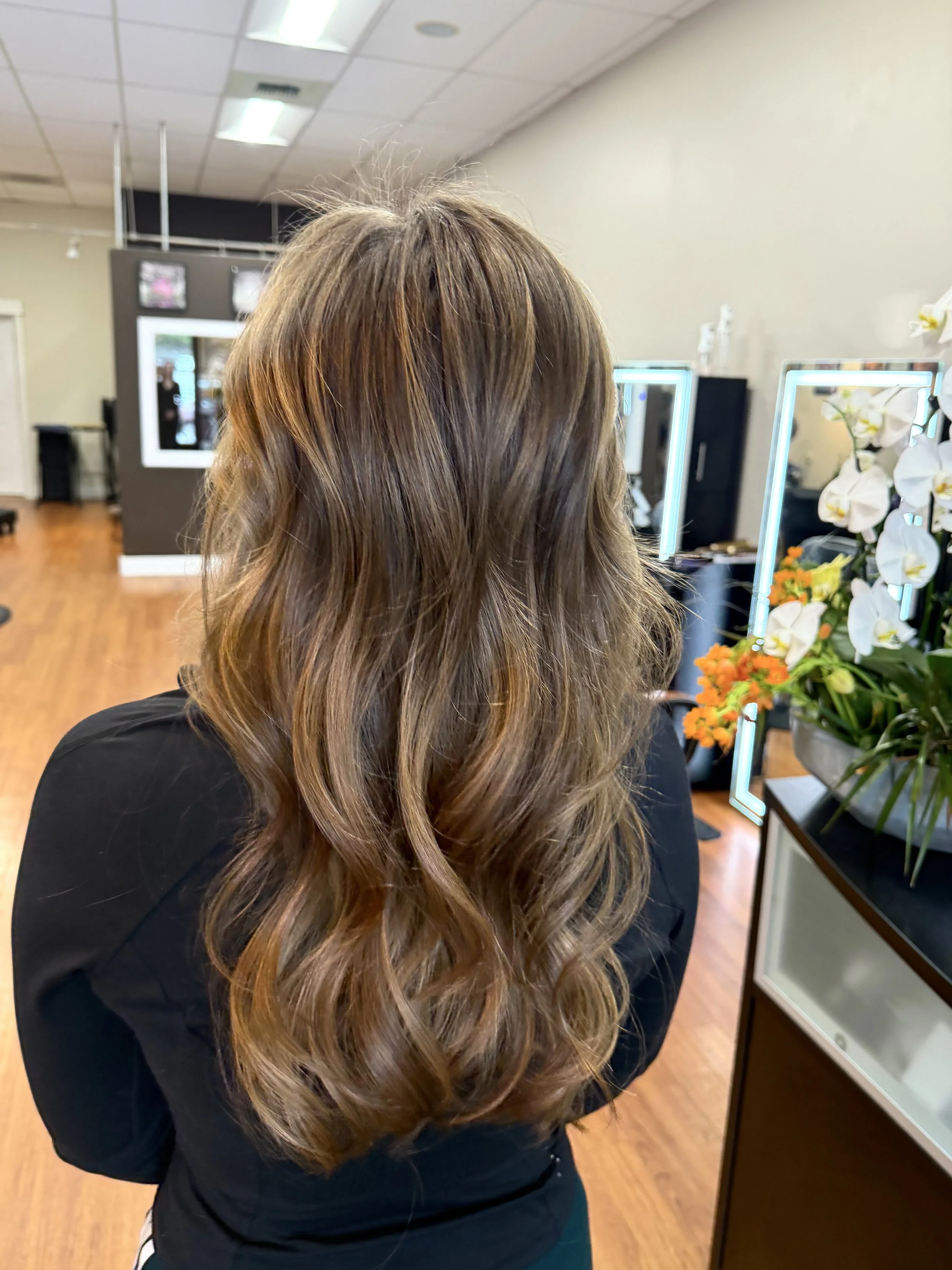 A person with long, styled brown hair standing in a salon or beauty studio with mirrors, flowers, and hair products visible in the background.