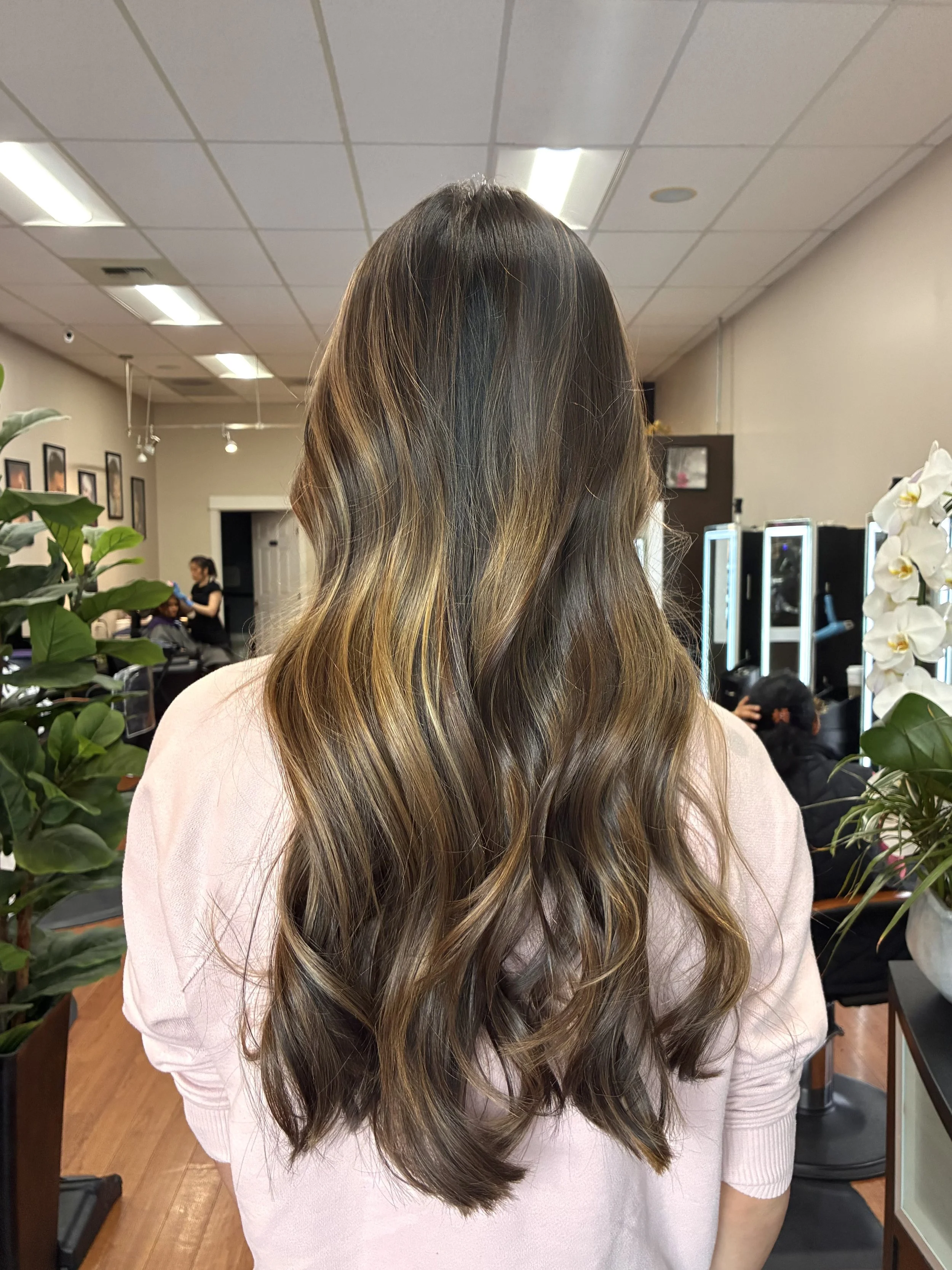 A woman with long, wavy hair featuring shades of brown and blonde standing in a salon, with salon equipment and other people visible in the background.