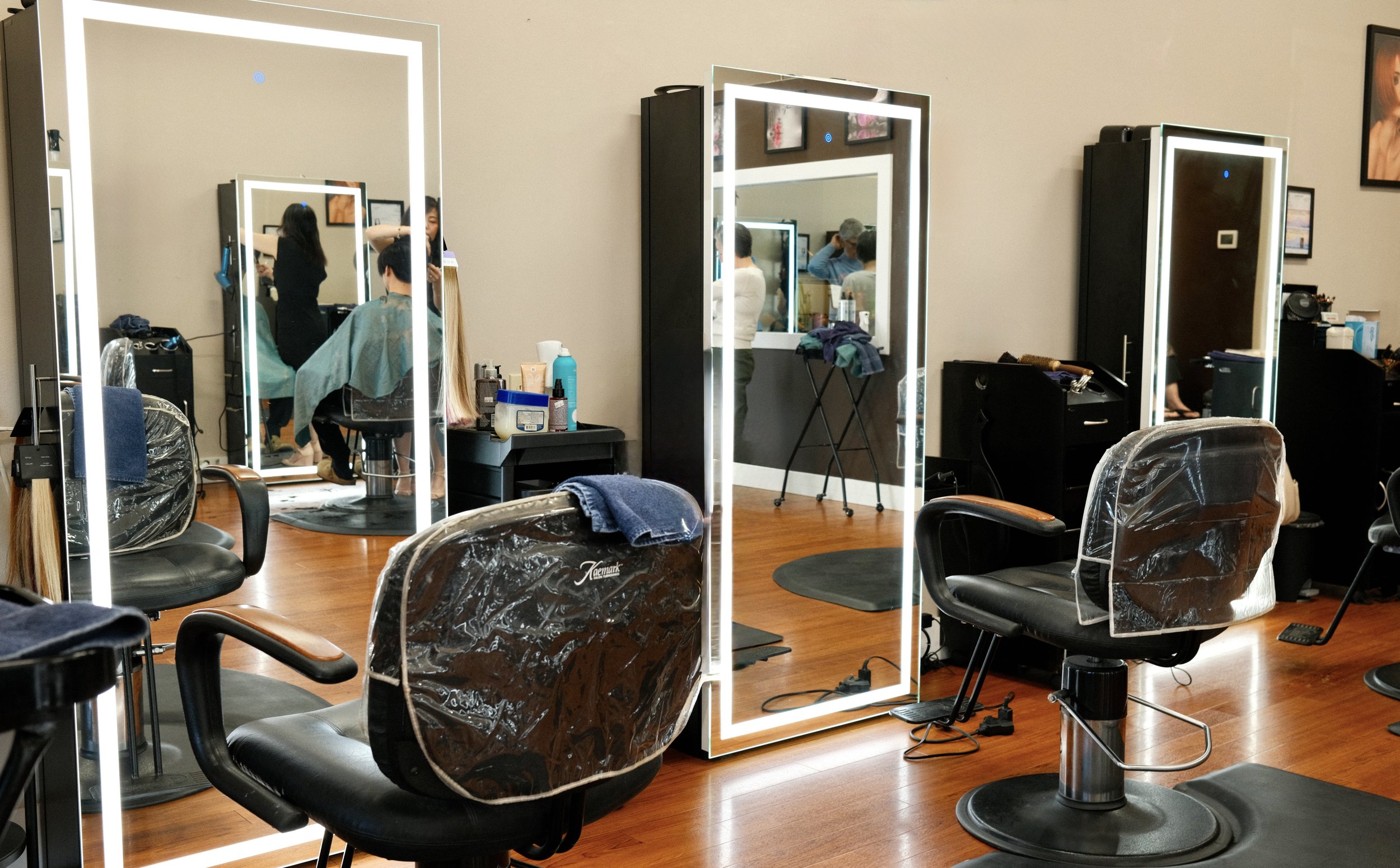 A hair salon with several salon chairs, mirrors with LED lighting, and hair styling tools. There are various supplies on counters and a couple of stylists working with clients.