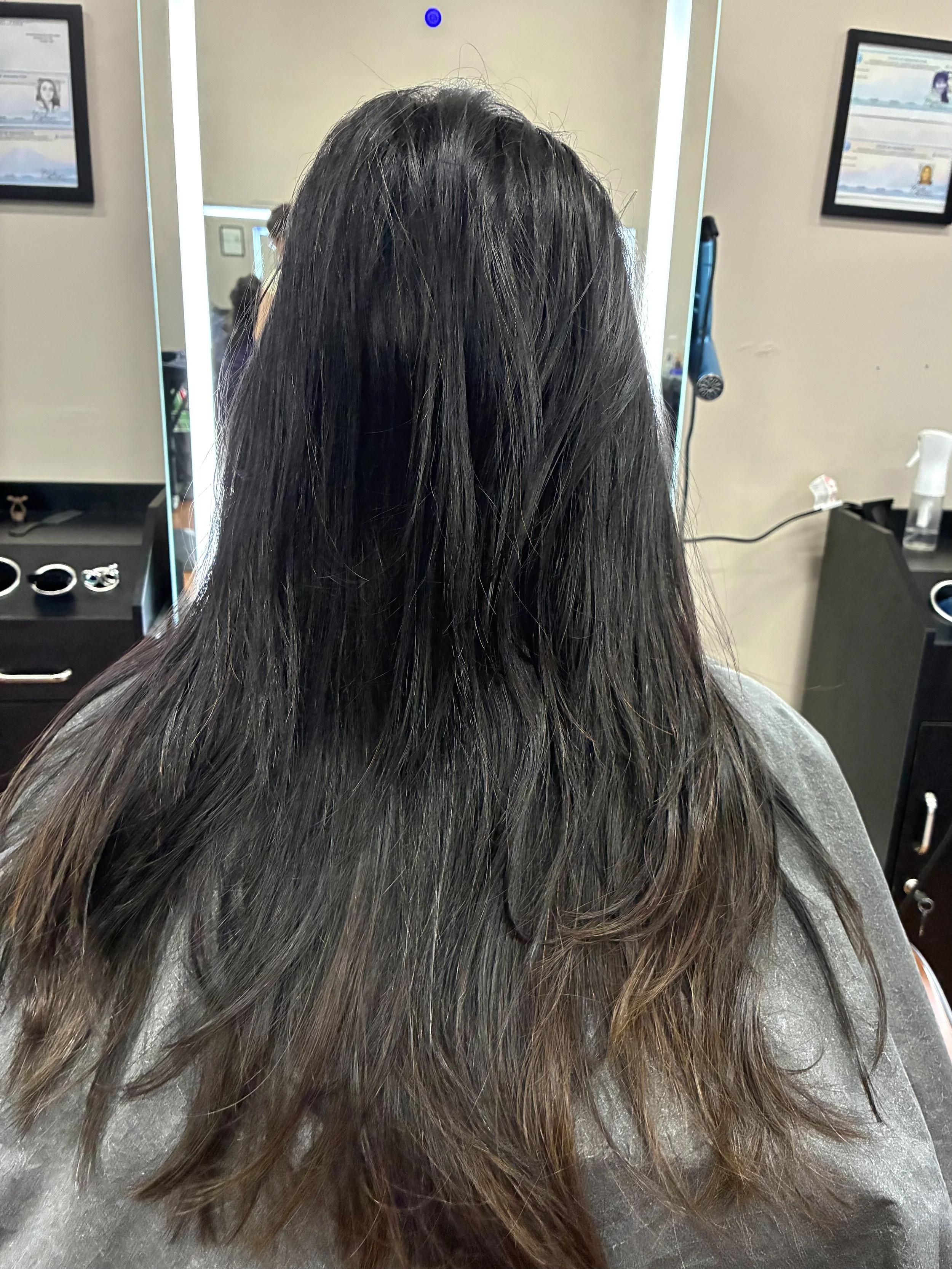 Back of a person with long dark hair sitting in a hair salon, with mirrors and salon tools visible in the background.