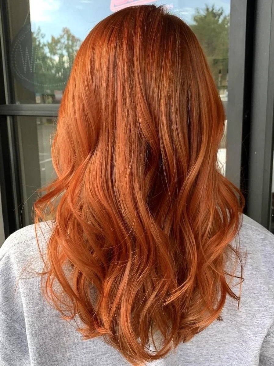 Back view of a person with long, wavy, vibrant red hair, wearing a light gray top, standing outside near a storefront window.