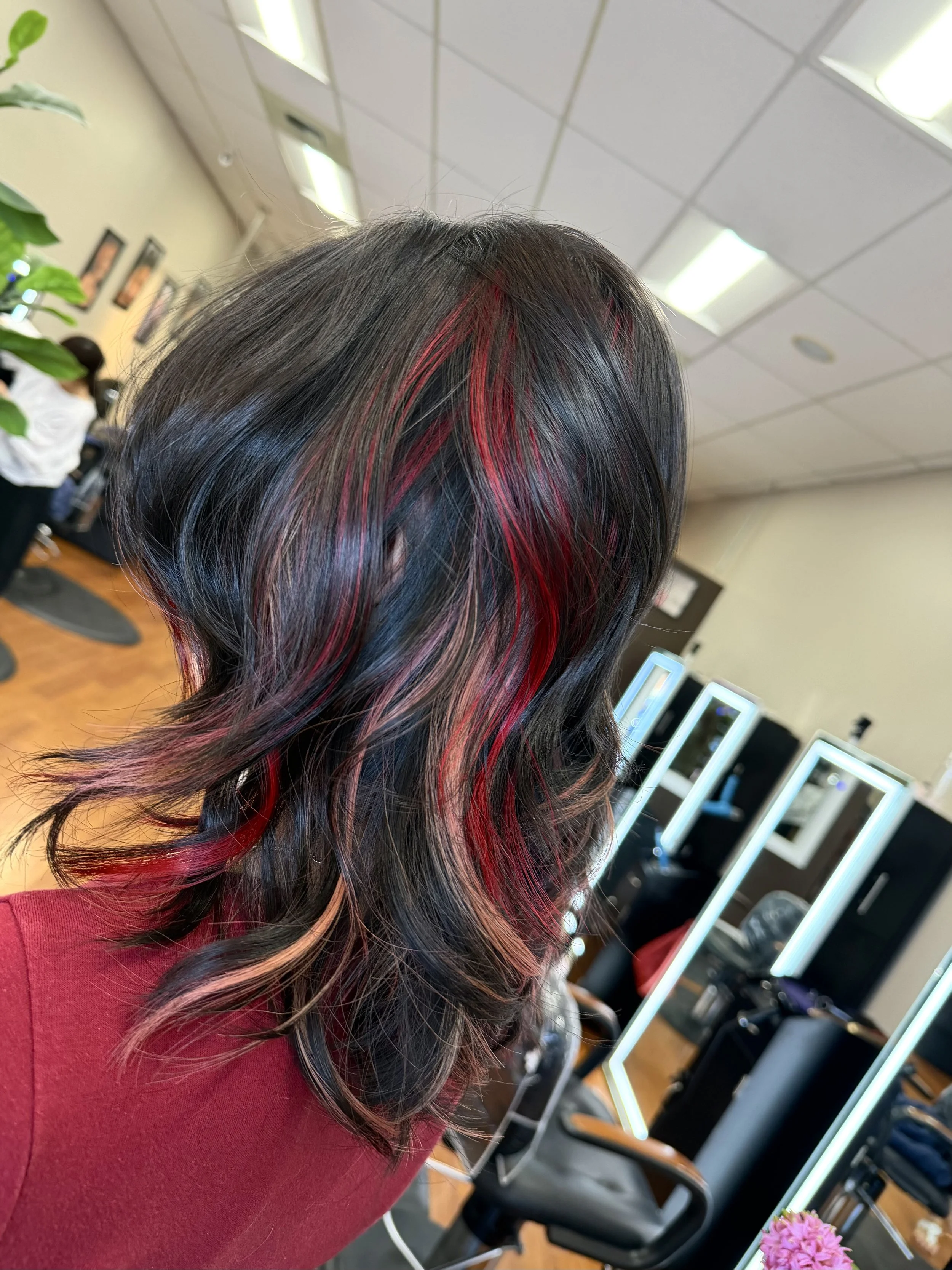 A woman with black hair and red highlights, wearing a red top, in a hair salon. The salon has bright lighting, mirrors, and other clients or staff in the background.