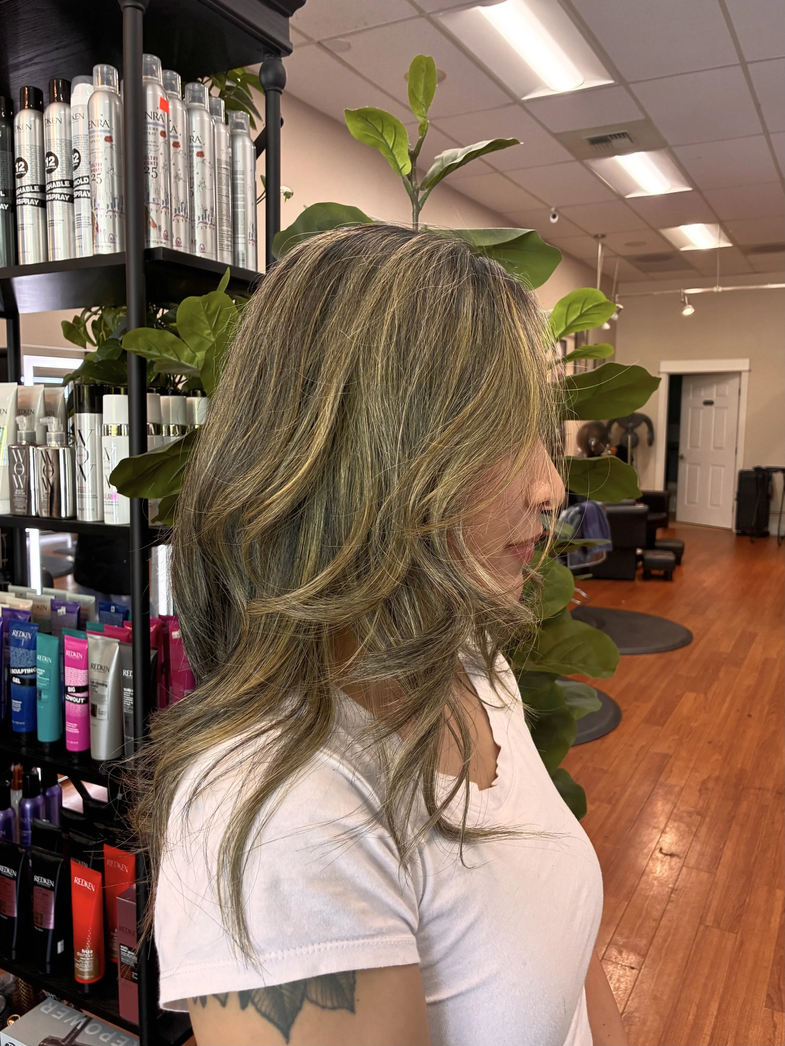A woman with long, wavy, blonde hair with subtle highlights stands in a hair salon. She is wearing a light-colored t-shirt and has a tattoo on her left arm. The salon has shelves with hair products, a large green plant, and a wooden floor, with brigh