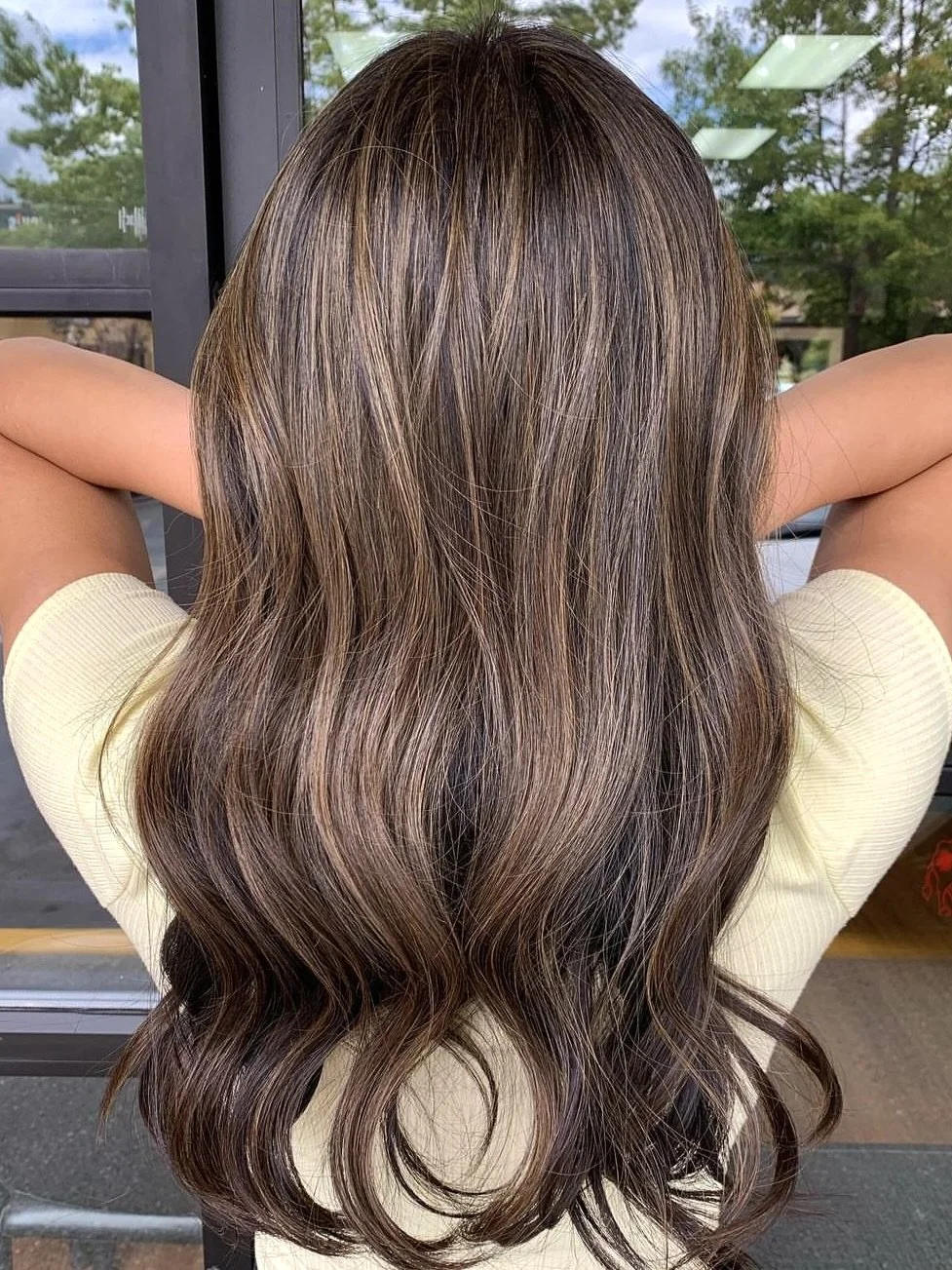 Back view of a woman with long, wavy, brown hair with highlights, standing near a door, with trees and sky visible outside.
