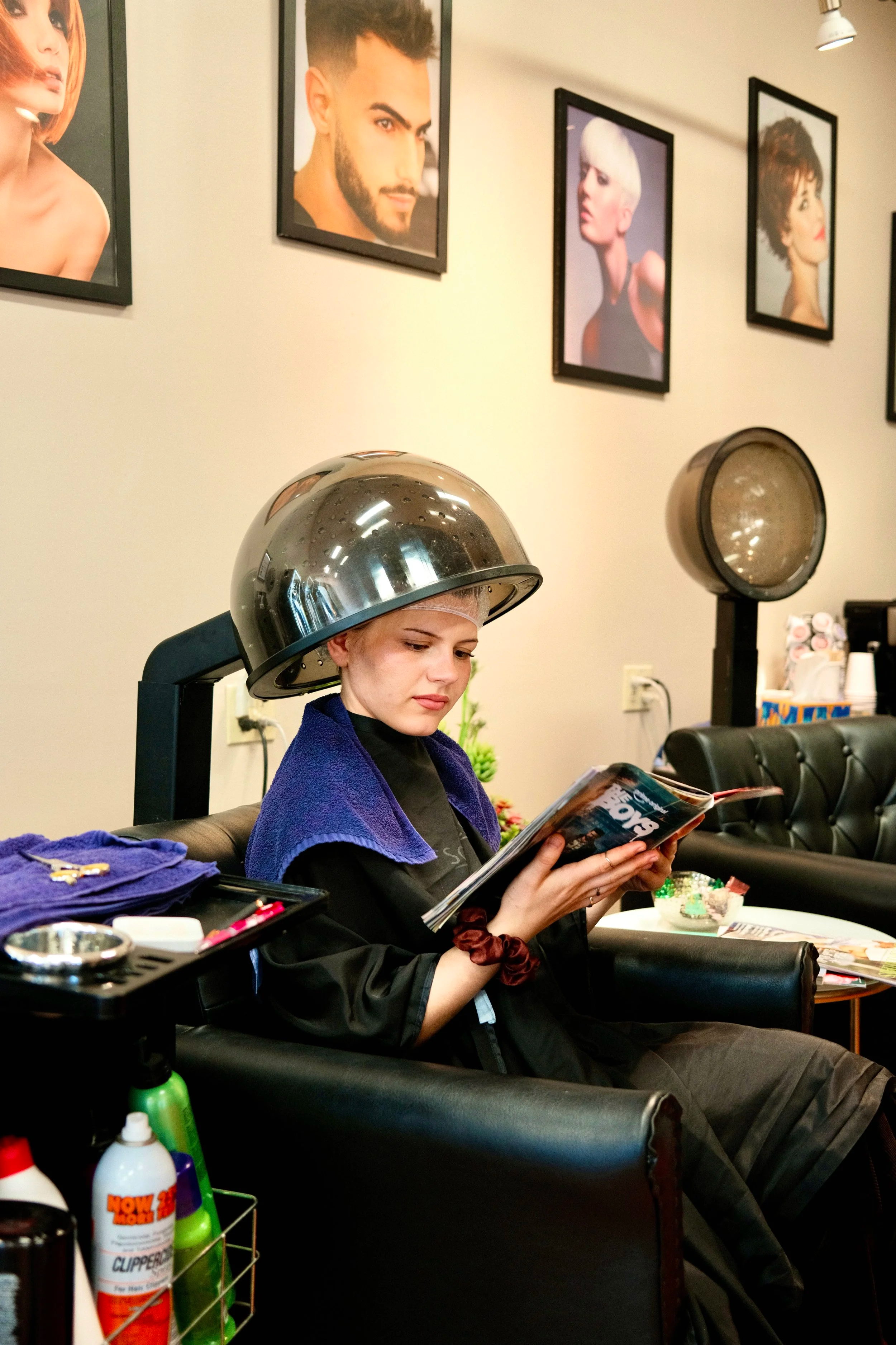 A woman with a hair dryer hood attached to hair dryer equipment, reading a magazine in a salon. The salon has framed hair portraits on the wall and hair styling supplies on a nearby table.