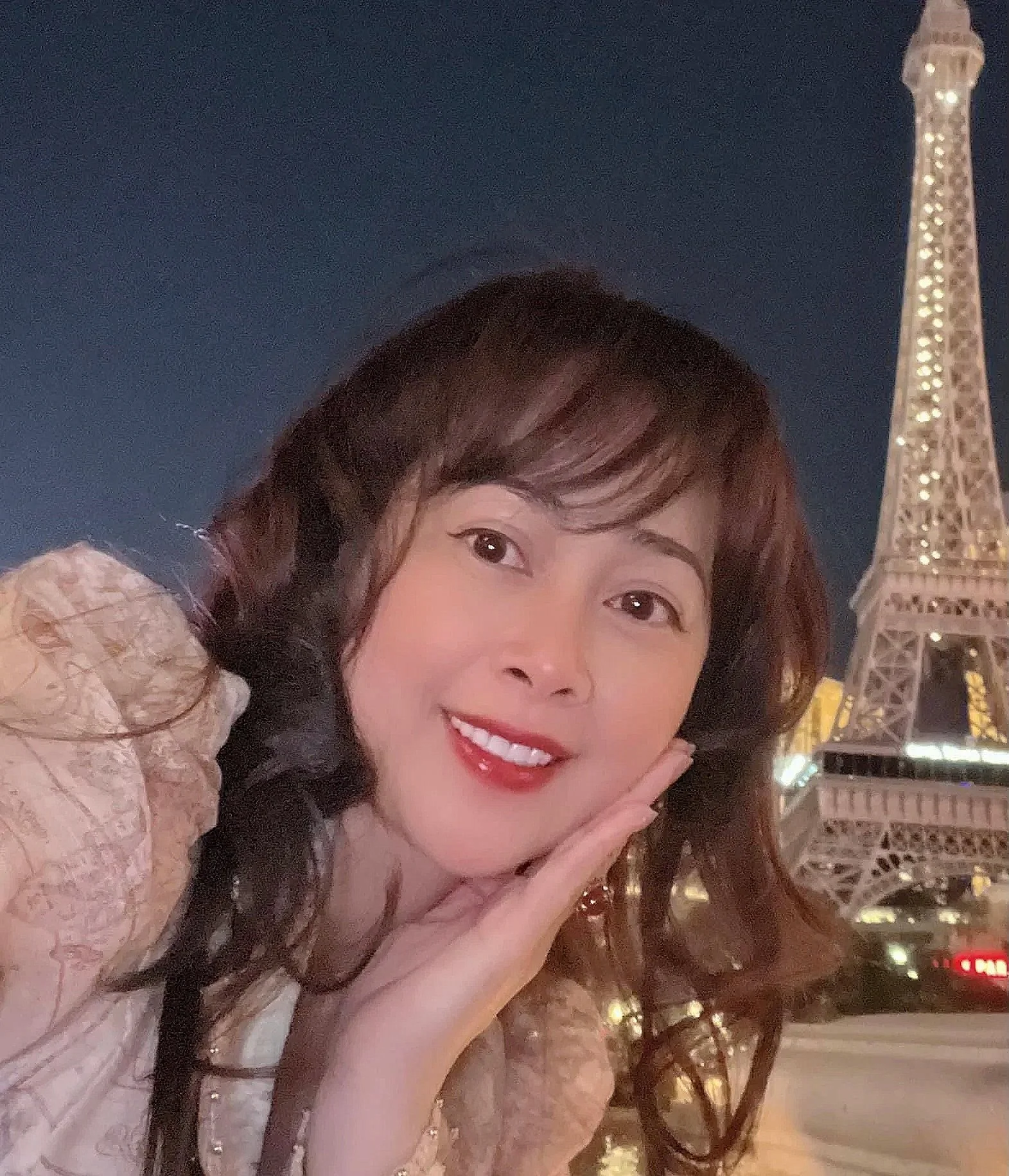 A woman with long wavy hair smiling with a finger touching her cheek in front of the Eiffel Tower at night.