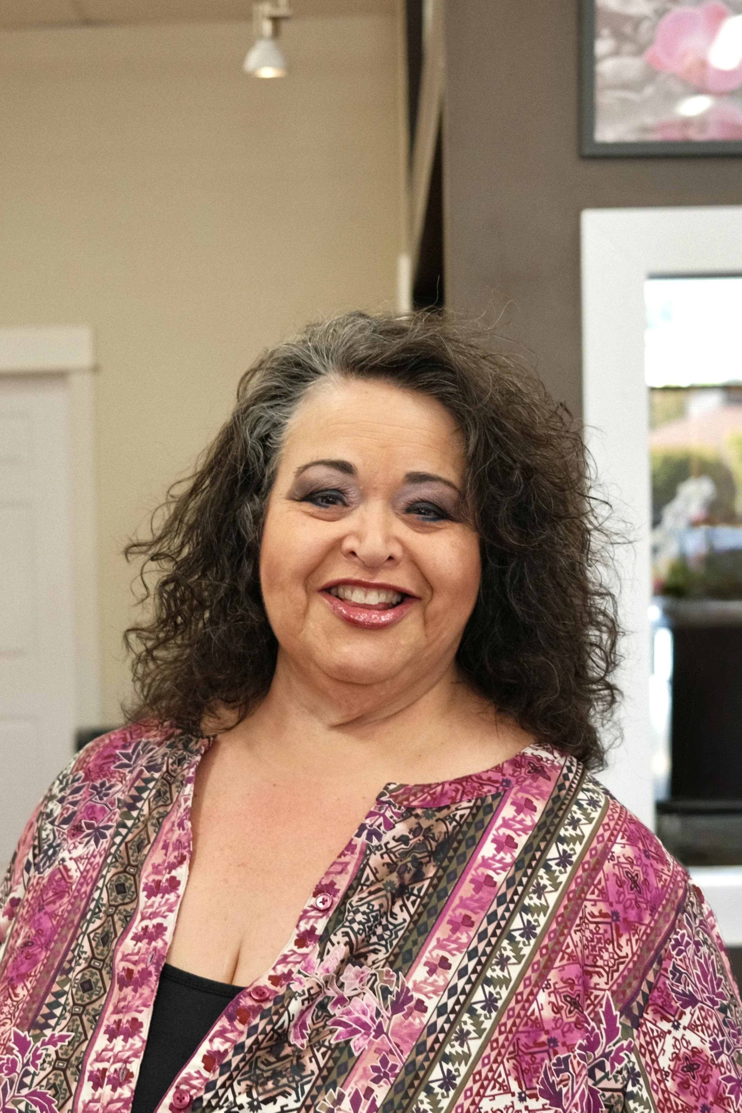 A smiling woman with curly gray hair wearing a patterned pink, purple, and black blouse inside a room with beige walls, a window, and a framed picture on the wall.