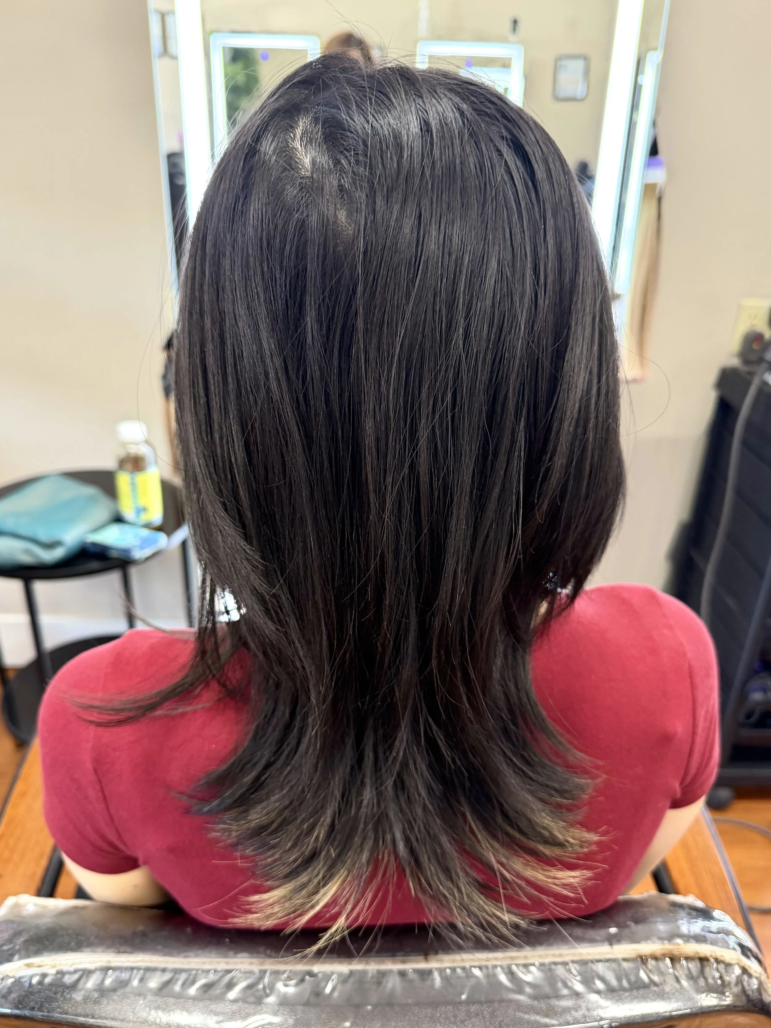 Person with long dark hair seated in front of mirrors in a salon, wearing a red shirt.