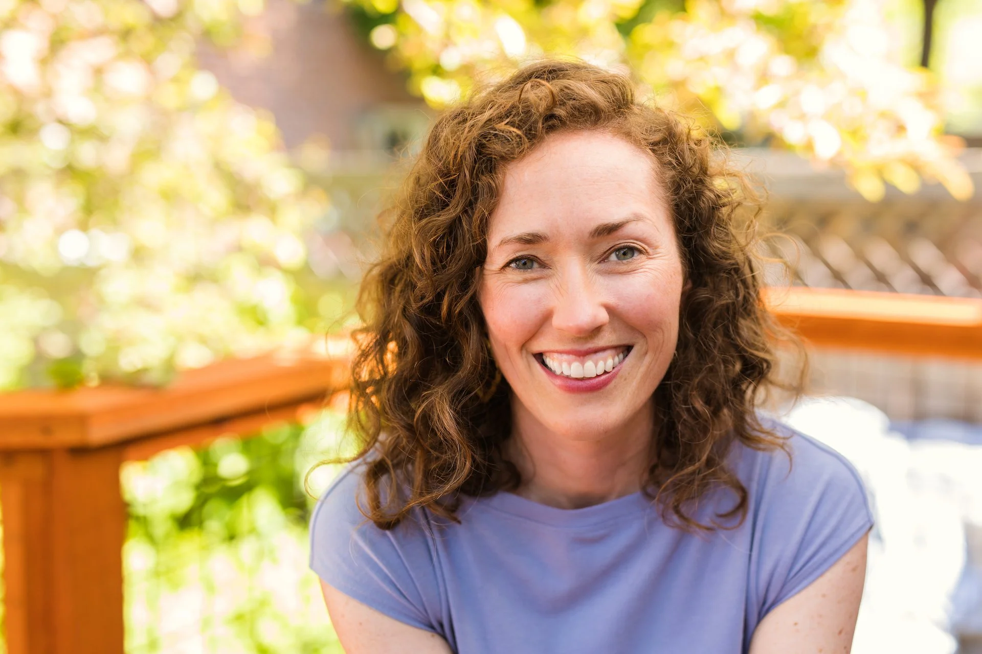 A woman with curly red hair and freckles smiling outdoors on a sunny day, with a blurred background featuring green trees and a wooden railing.