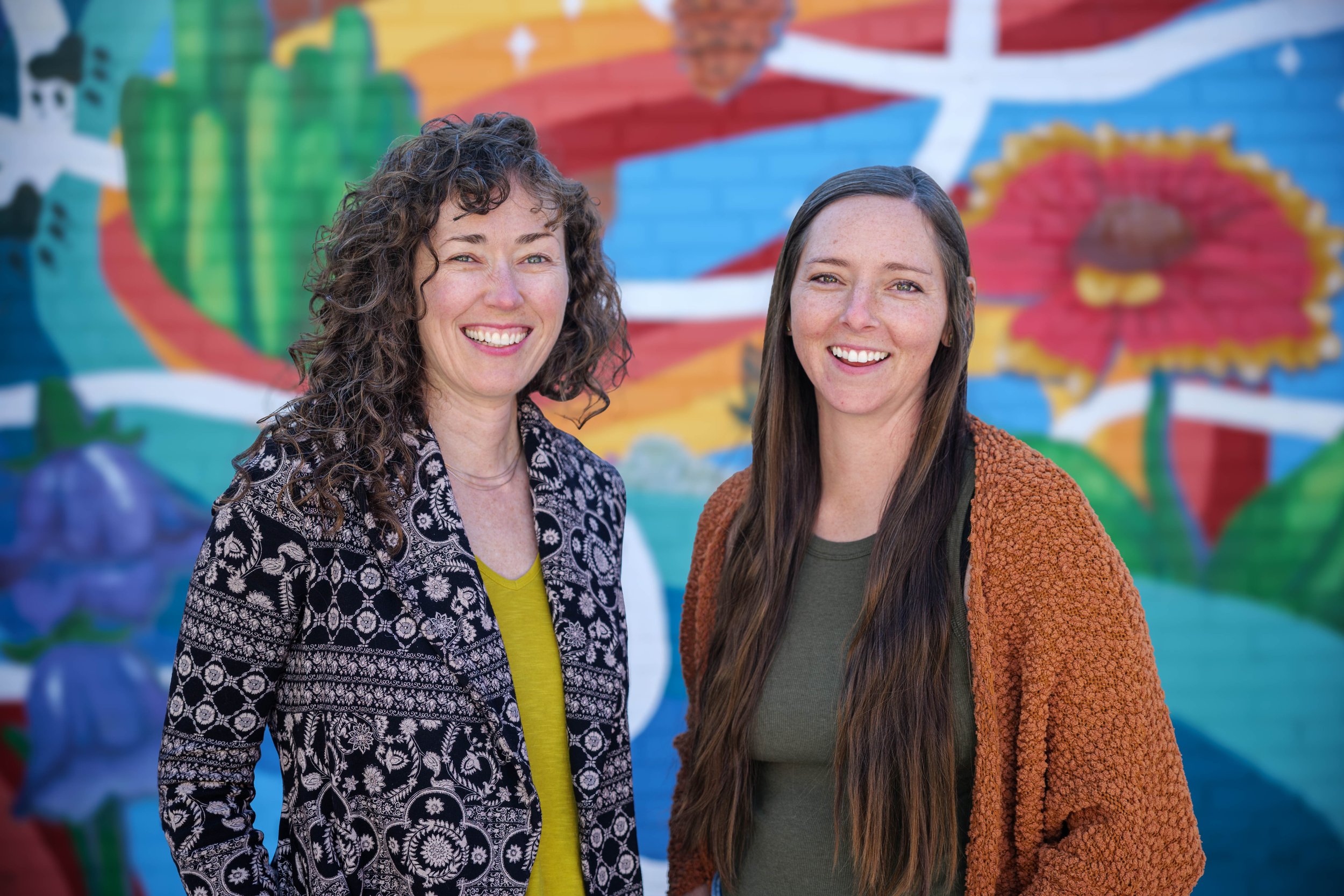 Two women smiling and standing in front of a colorful mural with birds and flowers.