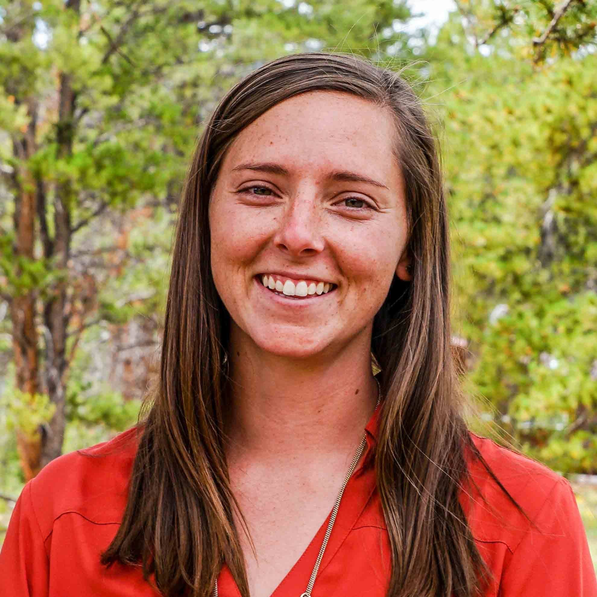 Close-up of a young woman smiling outdoors with trees and greenery in the background, wearing a red jacket.