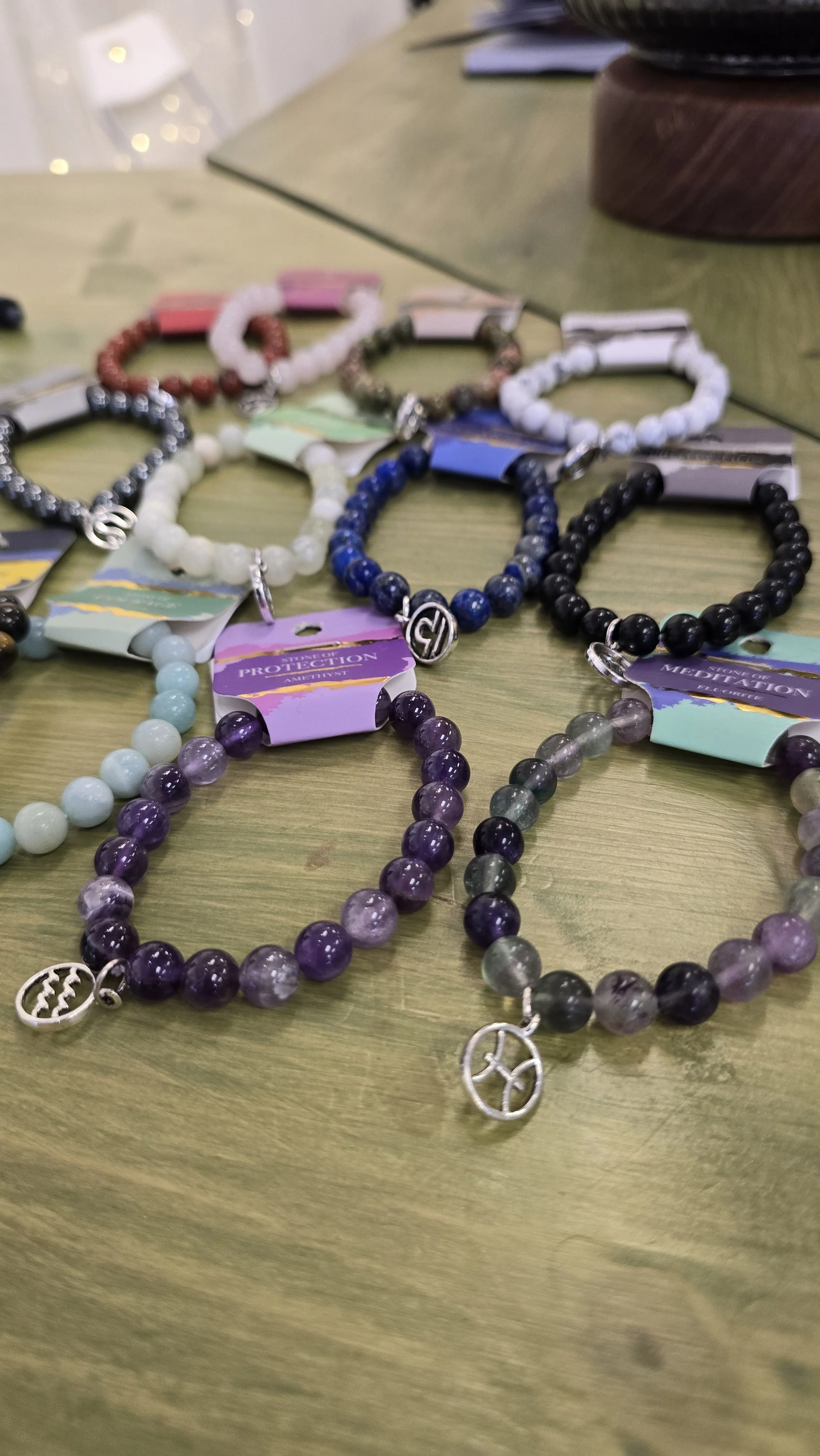 A collection of beaded bracelets displayed on a wooden table, featuring various colors including black, purple, white, red, and blue, with charms and tags attached.