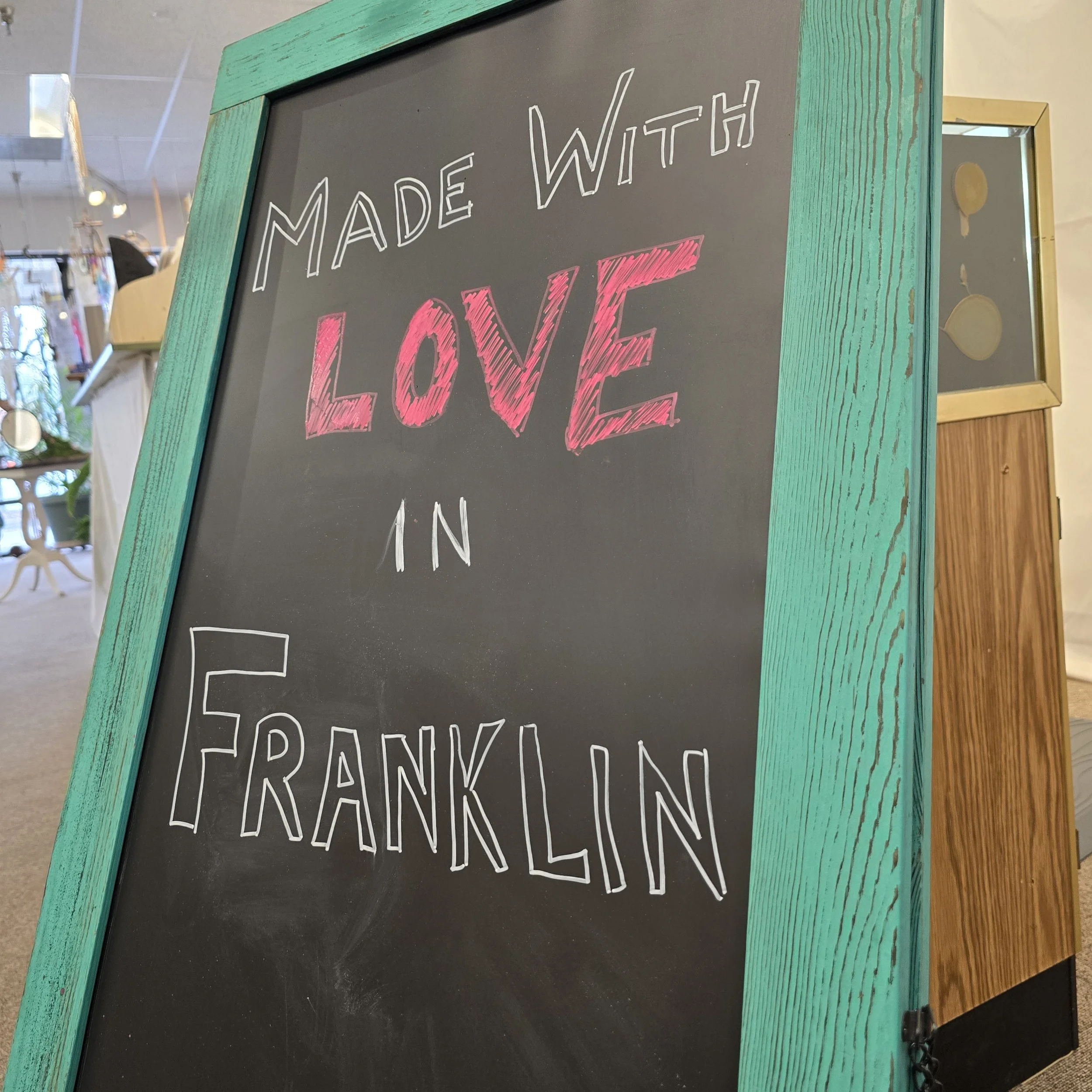 Black chalkboard sign with a turquoise wooden frame displaying the message 'Made with Love in Franklin' in white and pink chalk inside a store.