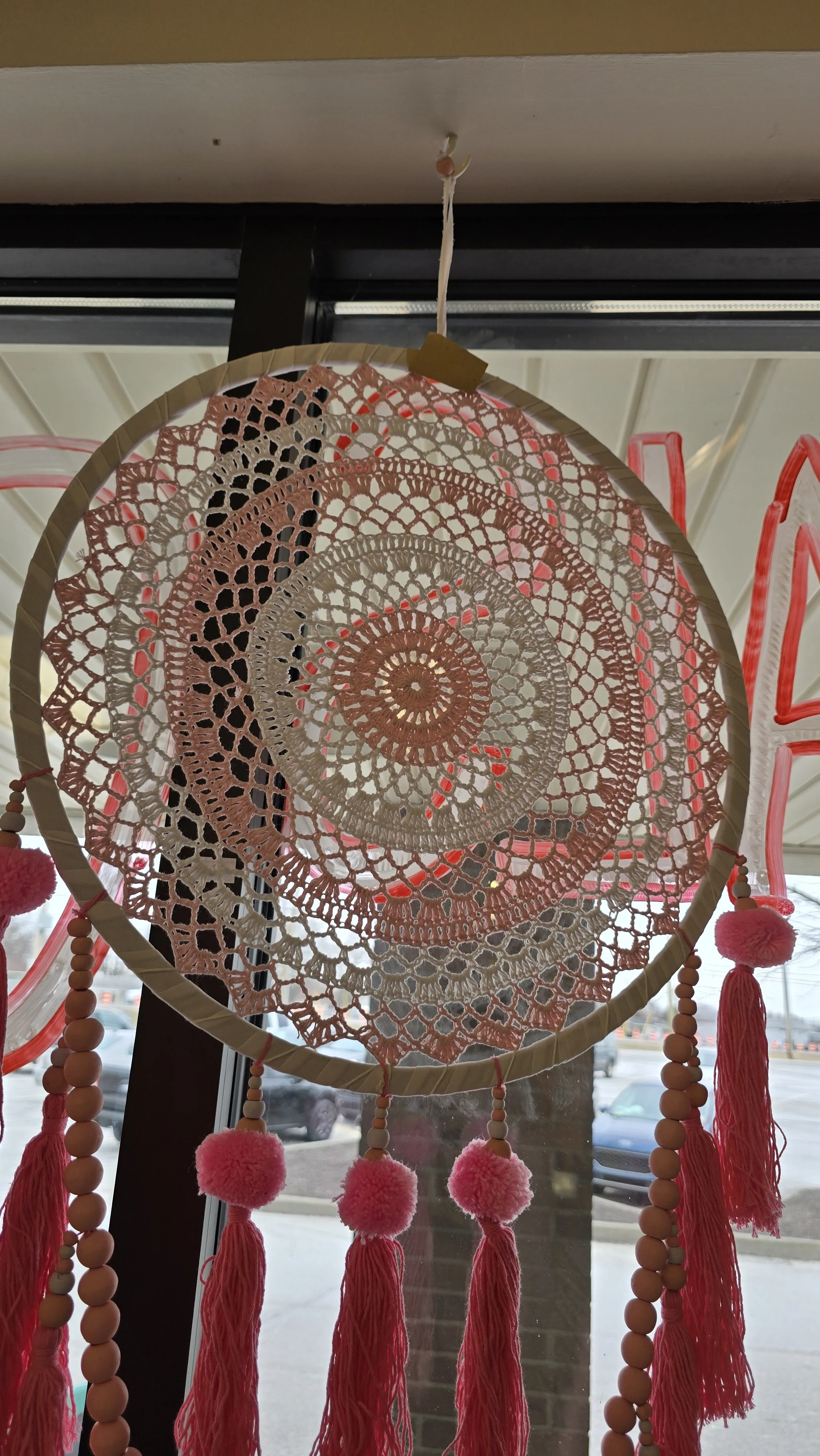 A pink and cream-colored dreamcatcher with crochet details, hanging from a window, decorated with pink pom-poms and wooden beads.