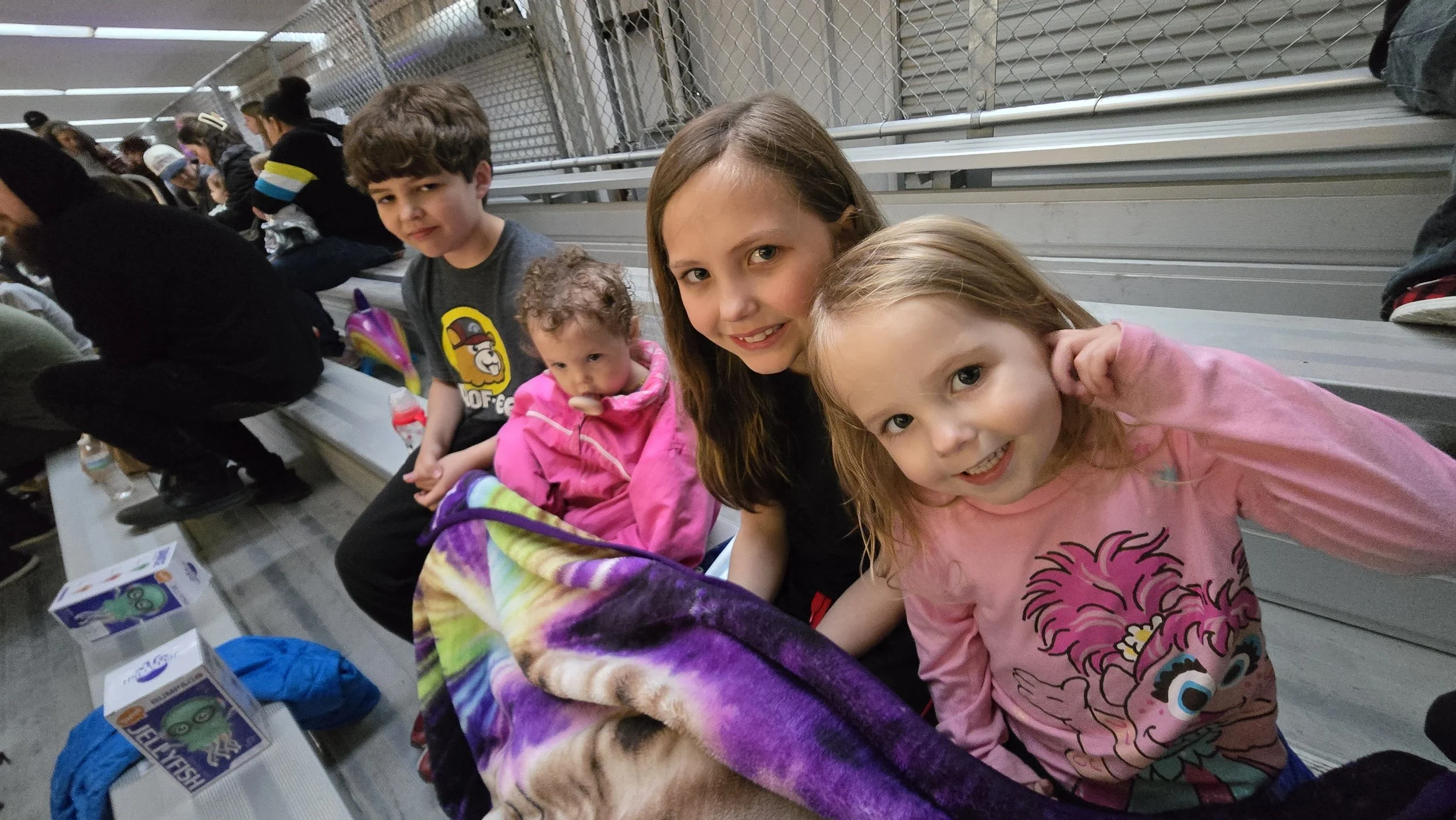 Group of children sitting on bleachers at an indoor venue, smiling and posing for the camera.