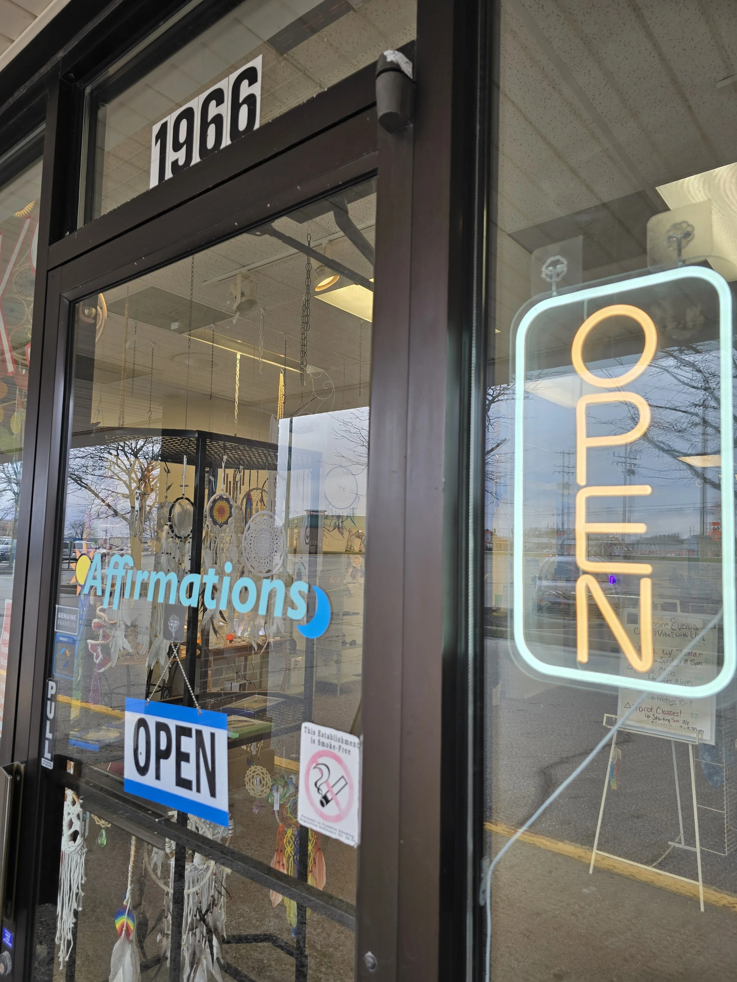 View of store entrance with large 'OPEN' neon sign, door window displaying merchandise like dreamcatchers, and signs indicating the store is open, with the year 1966 visible at the top of the door frame.