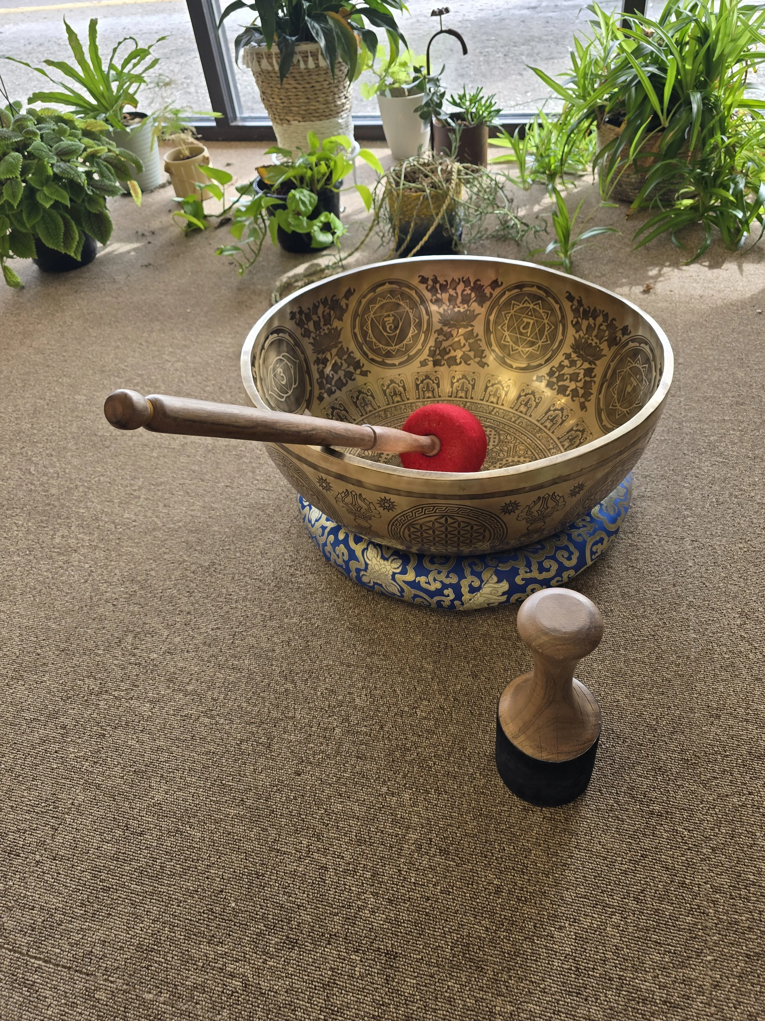 A large singing bowl with intricate gold patterns and symbols, a wooden mallet with a red felt ball, and several potted plants near a window with a carpeted floor.