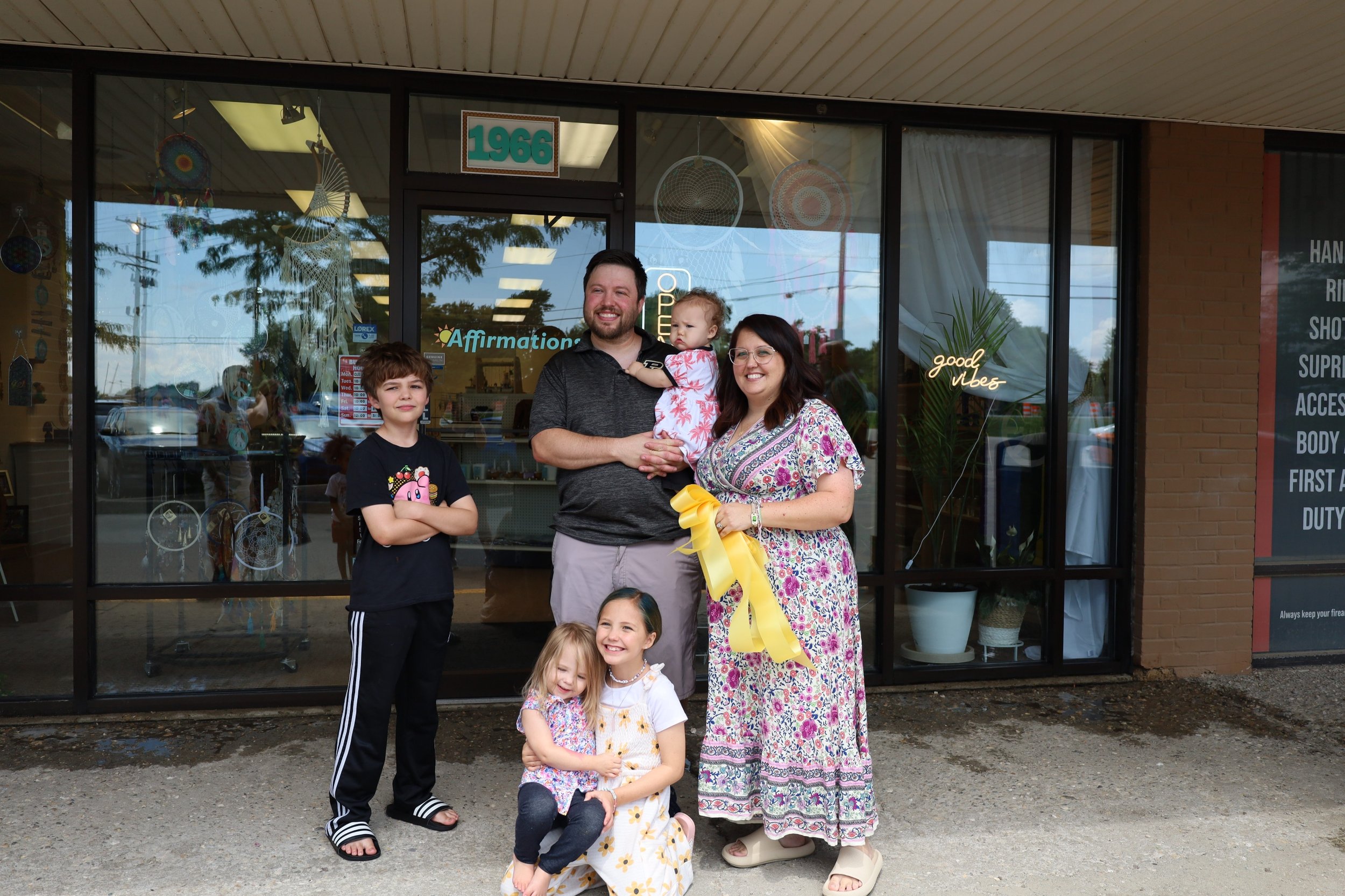A group of six people, including children and adults, standing outside a storefront. The group appears happy, with some smiling and one person holding a yellow ribbon. The storefront behind them has decorations and a neon sign that says "good vibes."