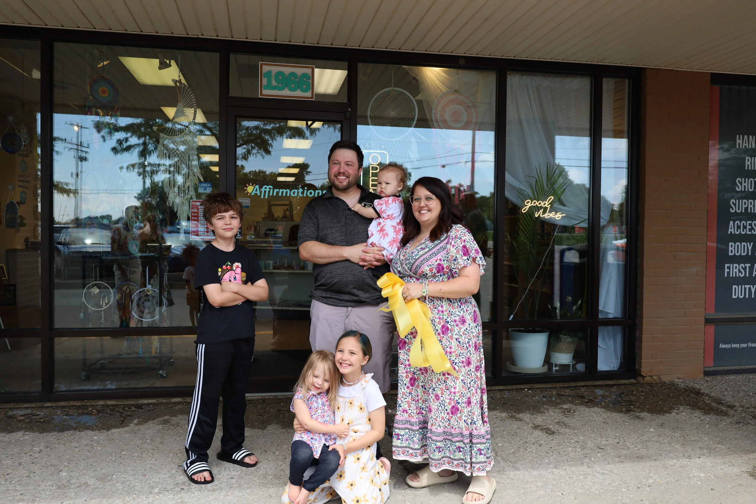 Family group of six standing outside a store with a glass storefront. The father holds a young girl in a pink and white patterned dress. The mother, wearing glasses and a floral dress, holds a yellow ribbon. Two girls and a boy are in front, smiling and posing for the photo.