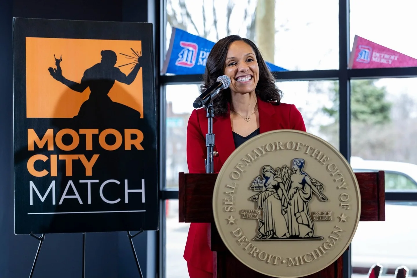 A woman in a red blazer standing at a podium with the seal of the City of Detroit, smiling and speaking into a microphone. Behind her is a sign that reads 'Motor City Match,' and windows showing an outdoor scene.