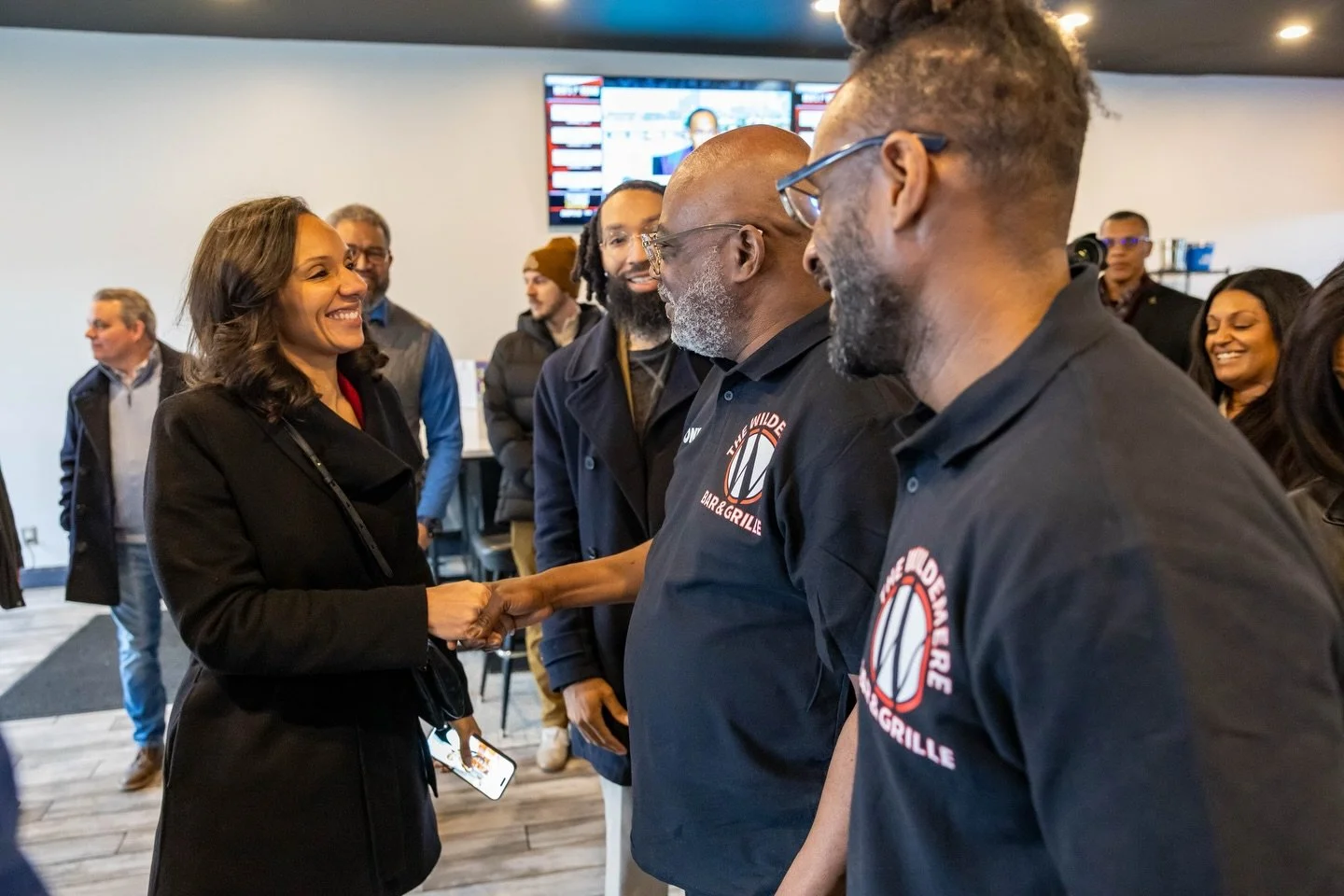 A woman in a black coat shaking hands with a man in a black T-shirt with 'The Wild Bar & Grille' logo, while other people gather around in an indoor setting.