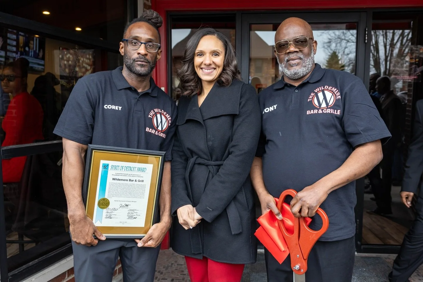 Three people standing outside a restaurant, two men and one woman, smiling. The man on the left is holding a framed award, and the man on the right is holding a red hydraulic bottle jack. They are wearing black shirts with the logo of The Wildemere B