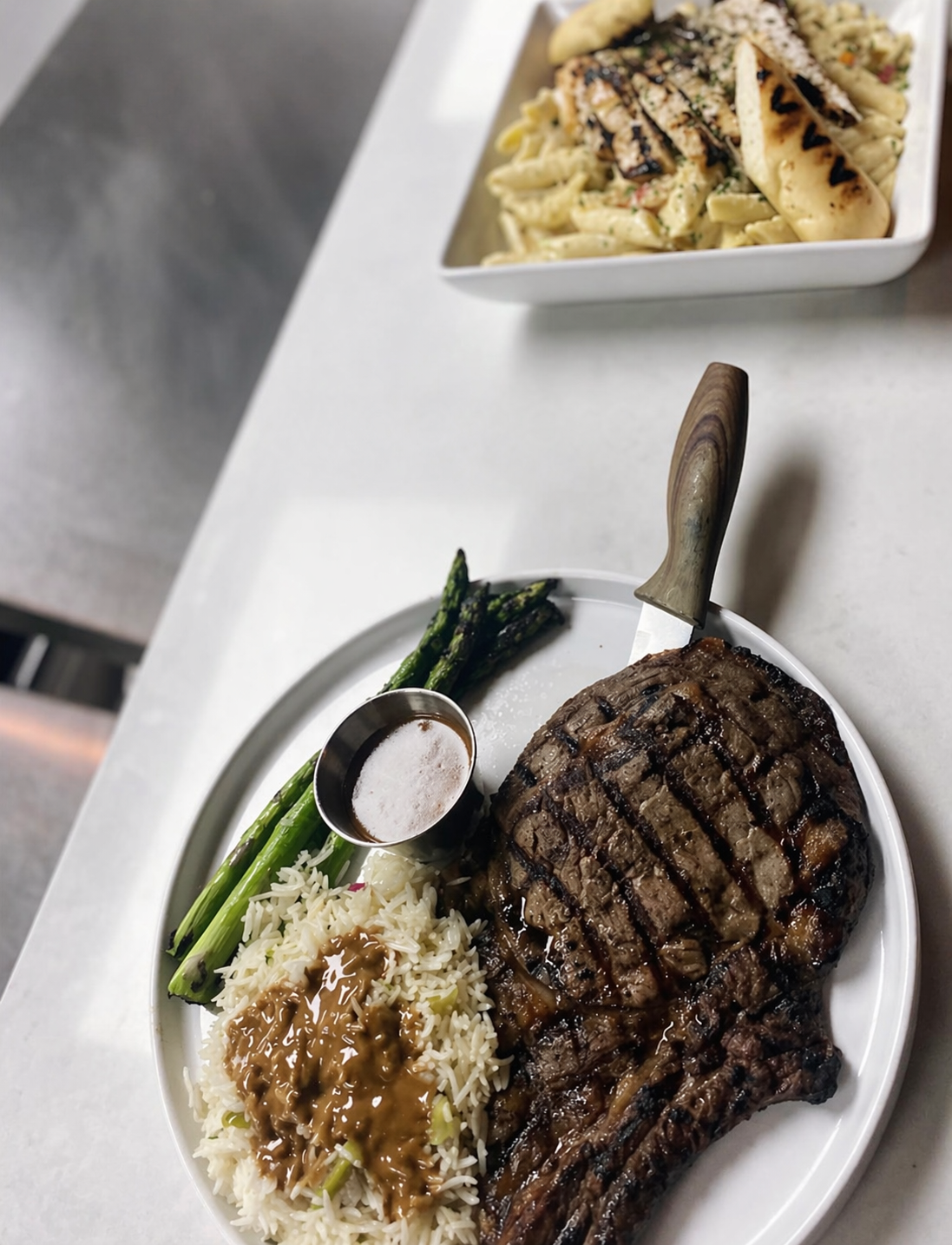 A plate of grilled steak with grilled asparagus, rice with sauce, and a small cup of sauce, on a white plate. In the background, a bowl of pasta with grilled chicken and herbs.